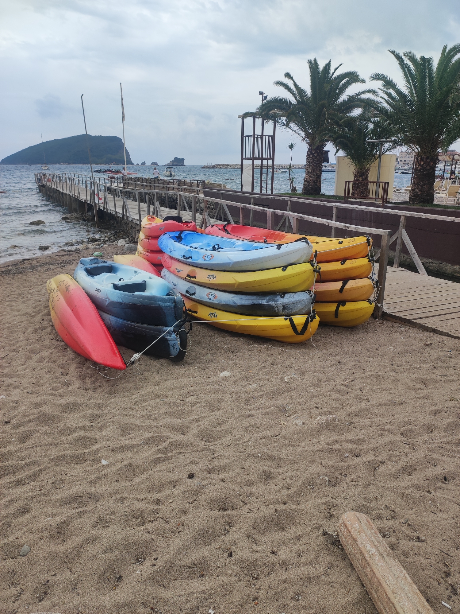 This image depicts a coastal scene on a cloudy day at a beach in Budva, Montenegro.
In the foreground, a sandy beach stretches across the bottom of the frame, showing subtle ripples and a piece of driftwood on the far right. Dominating the lower mid-ground are numerous kayaks, neatly stacked in groups on the sand. These kayaks are vibrant in color, including bright red, light blue, dark blue, and various shades of yellow and orange. Many of them bear the "RTM" brand logo, and several display numbers such as "29," "30," "31," and "28," suggesting they are rental or inventory items.
A long wooden pier extends into the sea from the left side of the frame, stretching towards a distant, large green island or hill. Two figures, likely men, are visible walking on the far end of this pier. Several small boats are scattered in the calm, grey-blue water near the pier and in the distance. Beyond the immediate coastline, smaller rocky islets can be seen in the sea.
To the right, behind the kayaks and parallel to the pier, a wooden fence or barrier marks the edge of a property. Behind this barrier, several tall palm trees rise, adding a tropical element to the landscape. A metal lifeguard stand with a ladder is partially visible behind the palm trees. Further in the background on the right, glimpses of beach lounge chairs and umbrellas suggest a resort or beach club area.
The sky is uniformly overcast with grey clouds, indicating a cloudy or possibly rainy day, yet the sea remains relatively calm. The scene conveys a tranquil, possibly off-season or early morning atmosphere, with the kayaks prepared for activity but no immediate signs of usage.