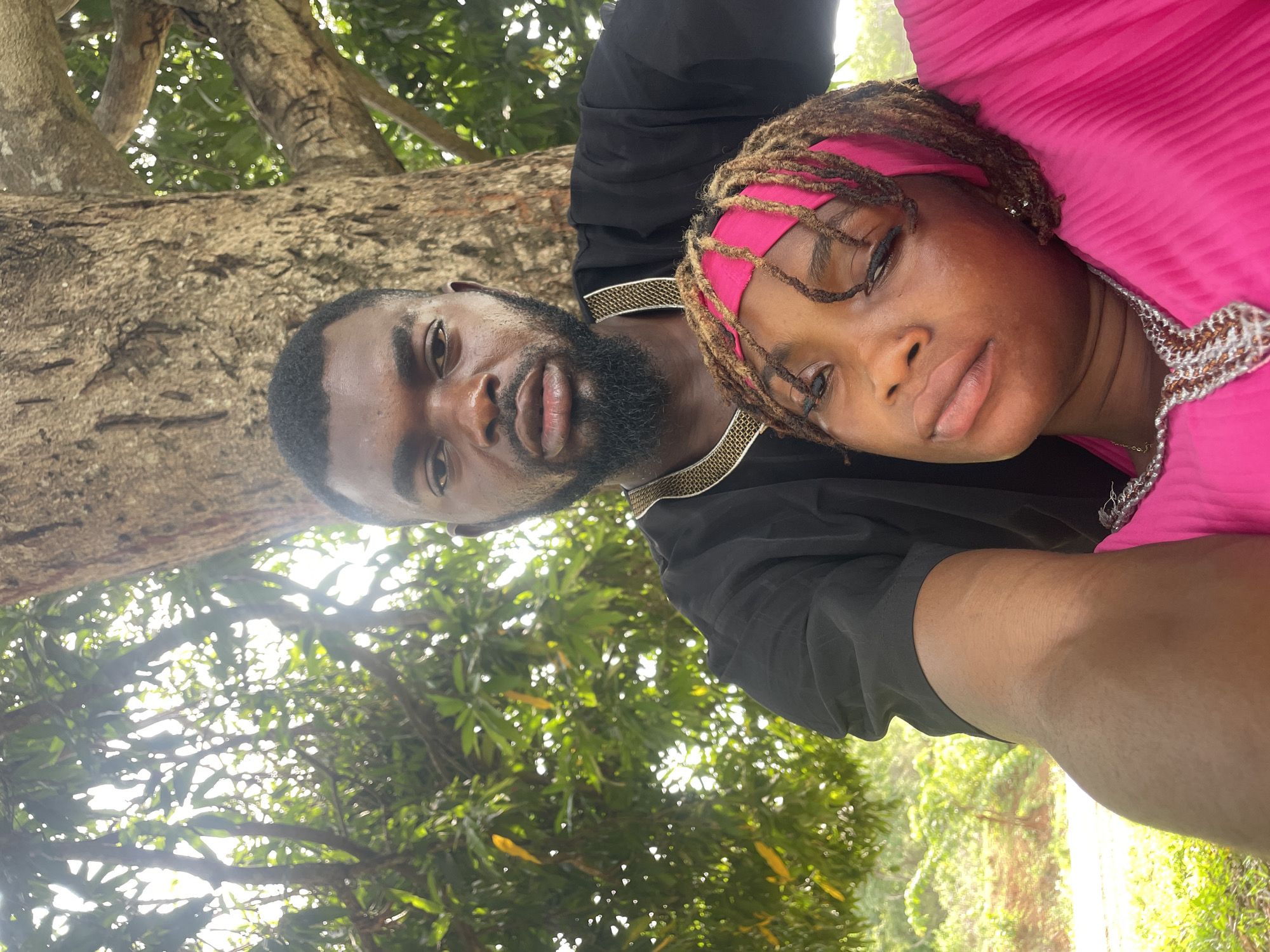 This image is a close-up selfie featuring two individuals, an adult male and an adult female, positioned horizontally against a natural outdoor backdrop in what appears to be Ogbe, Nigeria.
The male subject, situated on the left, has dark skin, a neatly trimmed beard, and short dark hair. He is wearing a black top adorned with an intricate gold and white patterned trim along the collar and shoulder line. His expression is neutral, and he looks directly towards the camera.
Beside him, the female subject also has dark skin and styled locs (dreadlocks), which are held back by a bright pink headband. She is dressed in a vibrant pink garment, possibly a top or dress, with some silver or white decorative elements near the neckline. Her eyes are partially closed, contributing to a relaxed or pensive demeanor as she also looks towards the camera.
The background is dominated by lush green foliage and the sturdy trunks of large trees, indicating a natural, possibly forested or park-like environment. Bright sunlight filters through the leaves, creating dappled light and suggesting a sunny daytime setting. No visible text is present in the image.