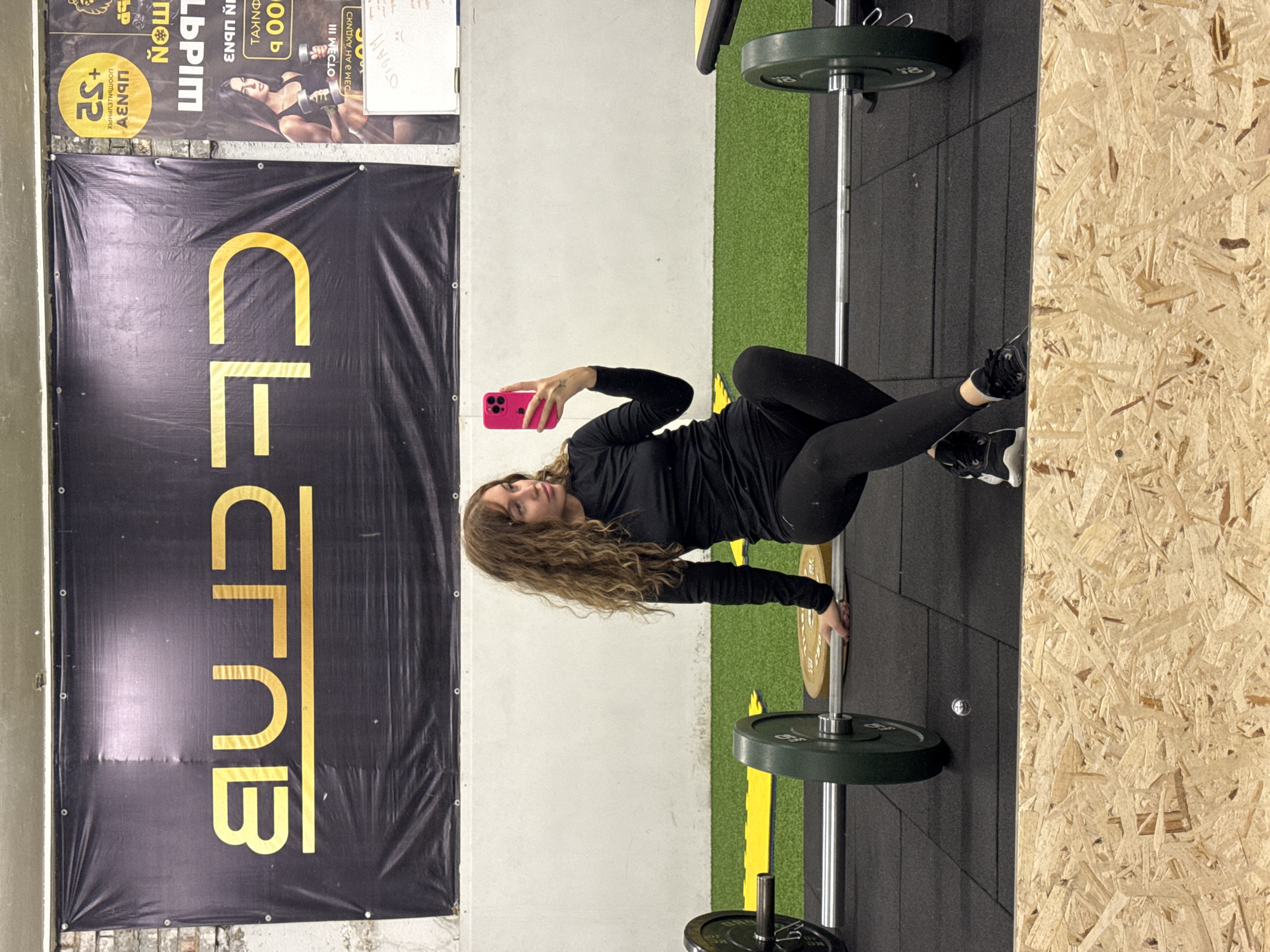 Here is a concise description of the image:
The image appears to be taken in a fitness center, possibly the "CF Club" based on the prominent banner in the background. A woman is positioned in the center of the frame, taking a selfie with a pink phone while seemingly in the middle of a workout. She is dressed in black workout attire. A barbell with weights is positioned near her, suggesting a weightlifting session. The background also reveals a section of artificial turf and a wall made of light-colored wood. Based on the text on the banner, it's likely located in the city of Chaikovsky, Russia.