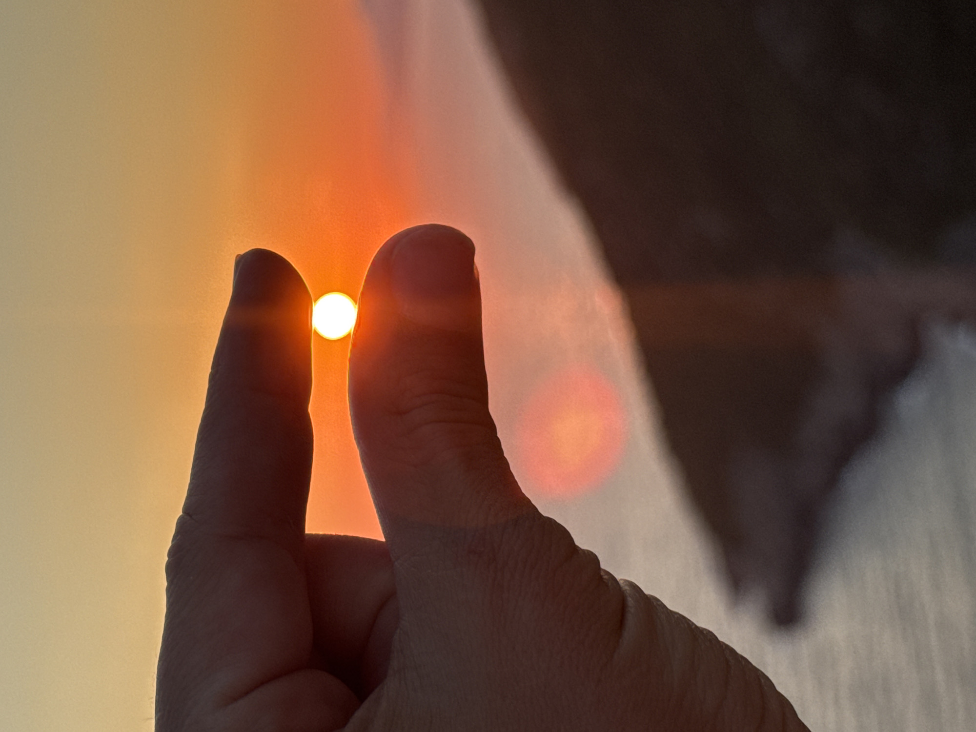 A person's hand, with fingers partially closed, is held up against a sunset. The sun appears as a small, bright orb nestled between the thumb and forefinger. The sky is a gradient of orange and yellow, typical of a sunset. In the background, out of focus, is a dark, silhouetted landmass that slopes down towards a body of water. The scene is likely captured at a coastal location, given the presence of water and landforms. The location is specified as Zagora, Montenegro.