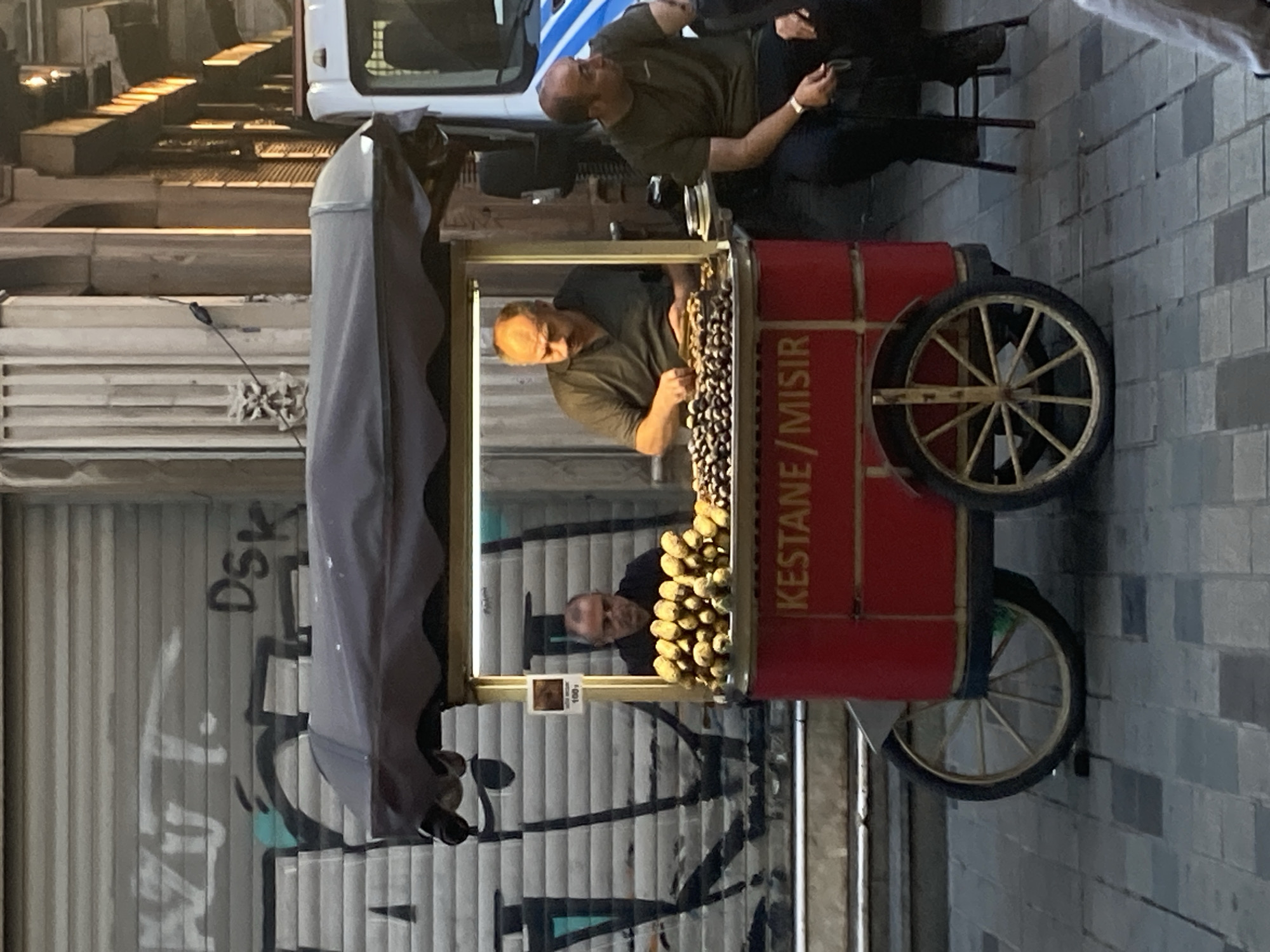 In Beyoğlu, Turkey, a street vendor operates a chestnut and corn cart on a paved sidewalk. The cart is red with a dark canopy and features the words "KESTANE / MISIR" (Chestnut / Corn) in white lettering. Two men are visible near the cart. One man, wearing a dark polo shirt, is seated to the right of the cart, holding a small object in his right hand. Another man, wearing a dark polo shirt, is behind the cart's serving window, handling food items. In the background, a metal roll-up door with graffiti covers a building facade, and a white vehicle is parked to the right of the cart. The scene is illuminated by overhead lights and ambient sunlight, casting shadows on the ground.