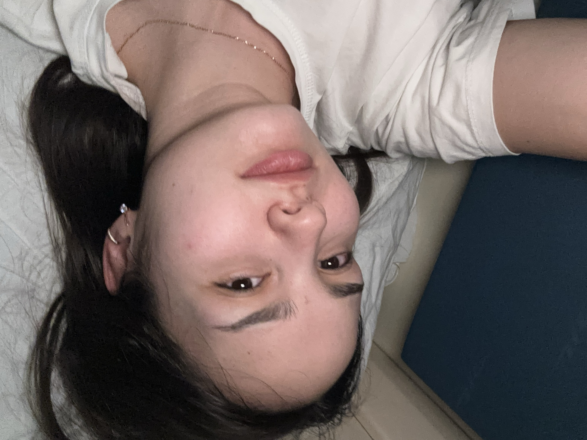 The scene takes place indoors in the city of Druzhinino, Russia. The image displays a close-up, angled perspective of an individual, identified as female, lying down. Her head is positioned towards the lower left of the frame, with her body extending towards the upper right. She possesses dark brown, long hair, which is spread across a light-colored, textured fabric surface, consistent with a pillow or bedsheet. Her eyes are open, dark brown, and directed slightly upwards. Her eyebrows are dark and well-defined, and her lips are slightly parted, revealing a natural pink hue. Her skin appears fair and generally unblemished. The individual is wearing a white short-sleeved t-shirt with a rounded neckline. A delicate gold-colored chain necklace is visible on her neck. Her left ear is adorned with a small silver-colored hoop earring in the helix and a small stud earring in the earlobe. A dark blue, textured surface is visible in the lower left foreground, while a beige wall or furniture component is partially visible in the upper left background. The ambient lighting appears dim. The individual is in a supine or reclined position, looking towards the point of capture.