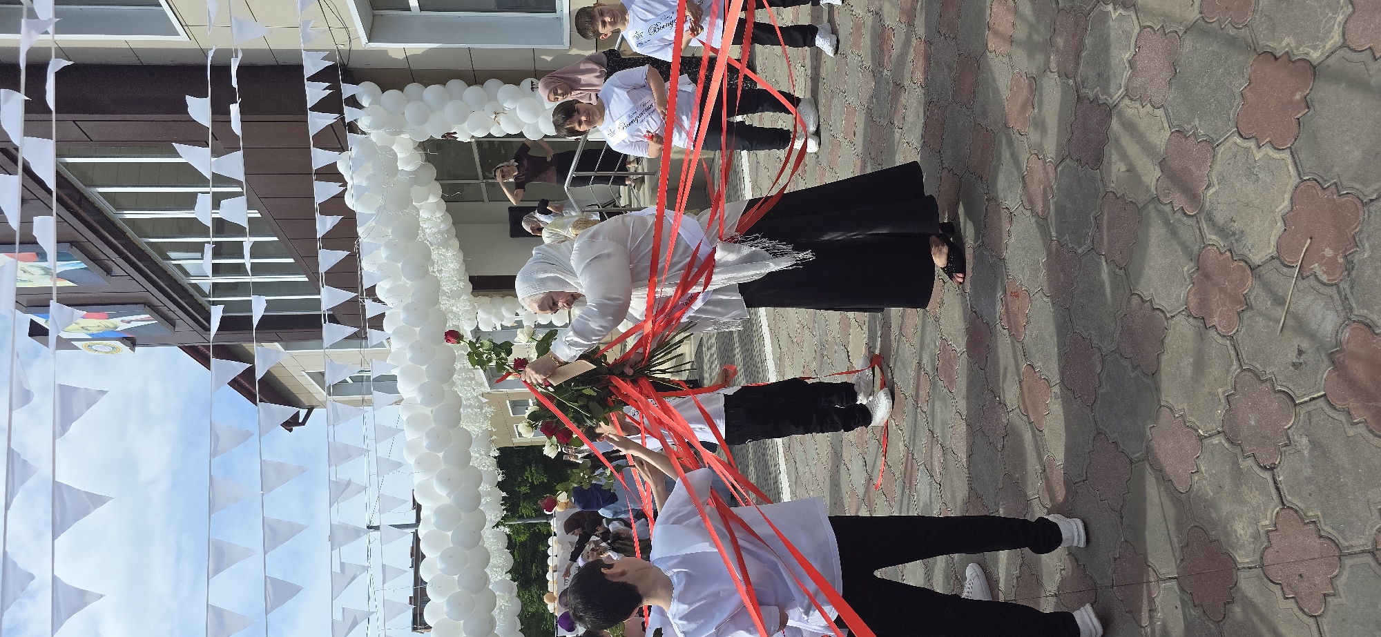 The image captures an outdoor scene in what appears to be a school or educational institution, located in the city of Unknown, Unknown. A group of people is present, most notably an elderly woman dressed in white clothing and a white headscarf, holding a bouquet of flowers that includes roses with red ribbons attached. There are also several students dressed in white shirts and black pants holding the ribbons. White balloons decorate the entrance of the building. White triangular flags are also strung up along the building's facade. The ground is covered in gray and red interlocking pavers. The sky is visible with some scattered white clouds.