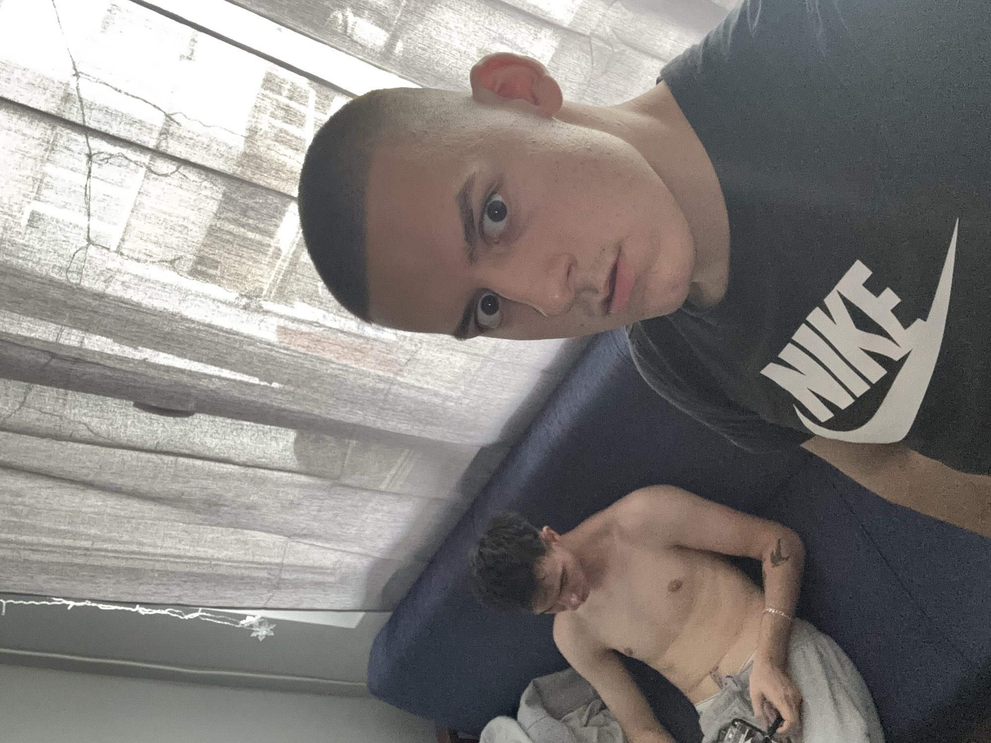 This is an indoor selfie taken at an oblique angle, featuring two young men in what appears to be a casual living space in Plovdiv, Bulgaria. The image is rotated, with a window covered by light-colored blinds visible in the background, suggesting natural daylight.
In the foreground, a young man with a short, buzz-cut hairstyle is taking the photo. He looks directly at the camera with wide eyes and a neutral to slightly intense expression. He is wearing a dark t-shirt with the white "NIKE" logo clearly visible on the front.
In the mid-ground, a second young man is relaxing on a dark blue sofa or bed. He is shirtless, revealing his torso, and is wearing light-colored shorts or sweatpants. He has dark, curly hair and is looking downwards, seemingly focused on an object he holds in his hands, possibly an electronic device or controller. A small tattoo, possibly of a bird, is visible on his left forearm, along with a bracelet on his left wrist.
The setting is an indoor room, likely a living room or bedroom, illuminated by soft, natural light filtering through the window blinds. The overall mood is relaxed and informal.