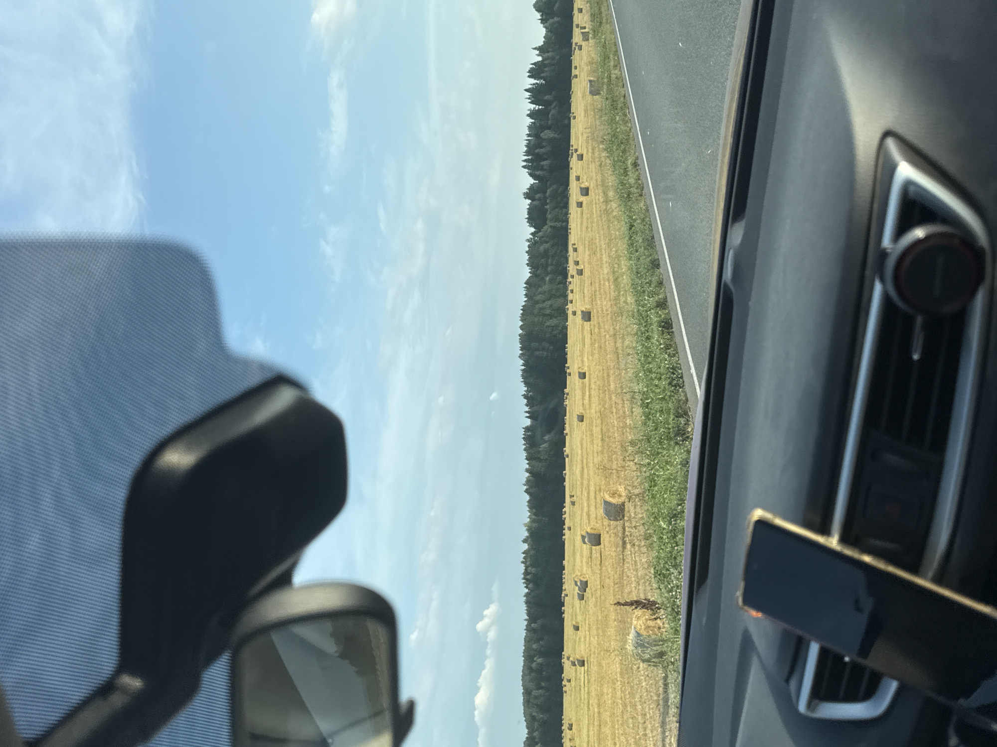 The image shows a view from inside a car, looking out at a rural landscape. In the foreground, the car's dashboard and a smartphone mounted on it are visible. Beyond the car, a field of hay bales stretches out, with a dense line of trees forming the horizon. The sky above is blue with scattered white clouds. The scene appears to be from a road trip in a rural area of Russia.
