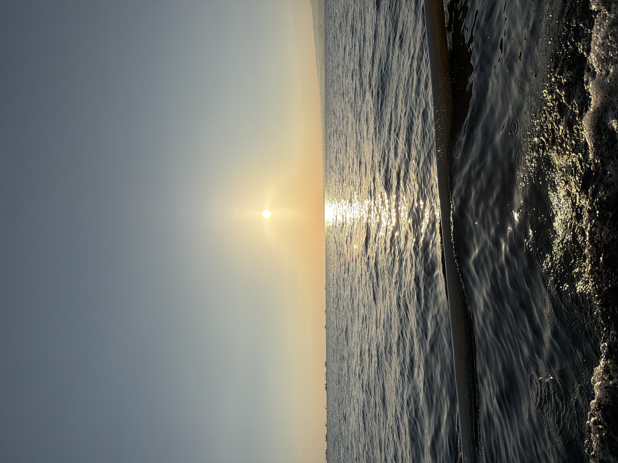 The image shows a sunset over the ocean. The sun is low on the horizon, casting a warm glow on the water. There are several boats visible in the distance. The water is choppy, with small waves breaking against the side of a boat in the foreground. The sky is clear and blue, with a few wispy clouds. The scene appears to be in the city of Unknown, Indonesia.