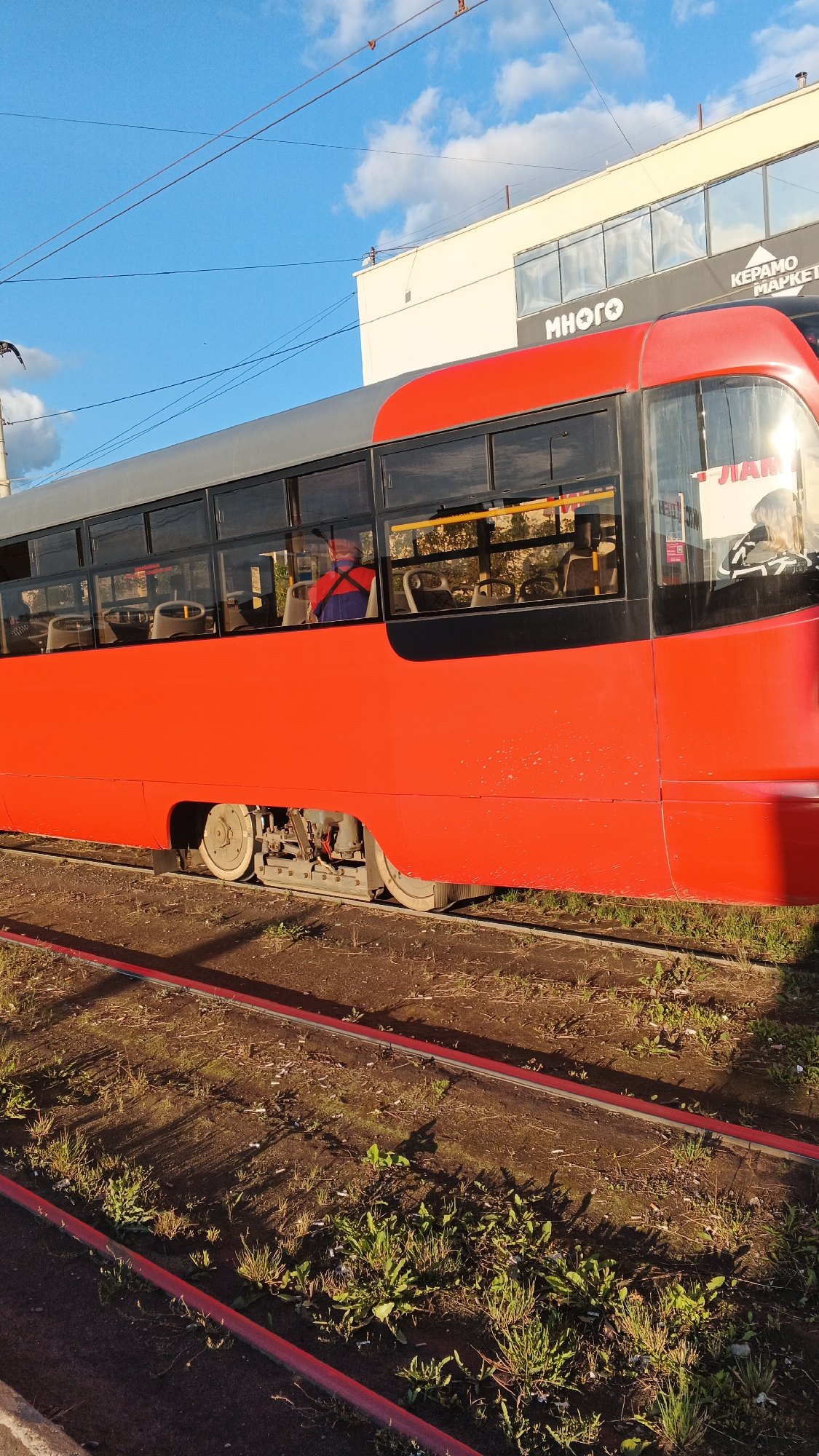 A red and gray tram is on tram tracks. The tram has many windows. Inside the tram, a person wearing a blue and red shirt is visible. In the background, there is a building with "МНОГО" (which means "many" in Russian) and "КЕРАМО МАРКЕТ" (which means "Ceramic Market" in Russian) written on it. The scene is outdoors with a blue sky and some clouds. The tram tracks are surrounded by dirt and grass. The image appears to be taken during daylight, possibly late afternoon given the angle of the sun. This scene is likely in Izhevsk, Russia, based on the Russian text on the building.