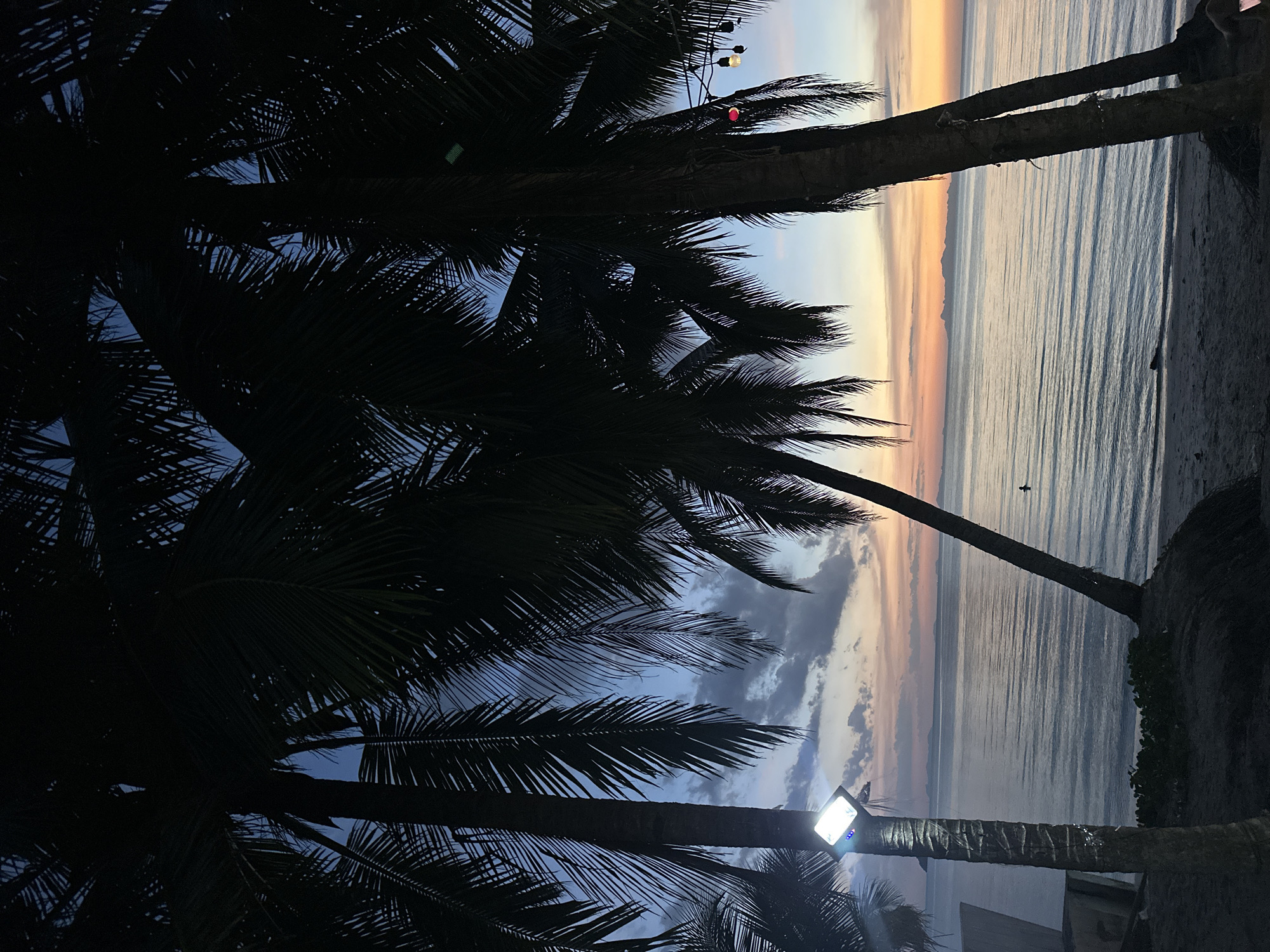 The image captures a twilight scene on a beach in Ko Samui District, Thailand. In the foreground, silhouetted palm trees frame the view. A bright, square solar-powered light is attached to one of the palm trees on the left. The calm ocean stretches to the horizon, reflecting the pastel colors of the fading sunset. In the distance, a faint outline of islands can be seen. A lone figure is visible in the water, appearing small against the vastness of the sea. String lights with two bulbs, one appearing red and the other white, are also present, hanging among the palm fronds on the right side of the frame. The sand of the beach is dark and textured in the foreground.