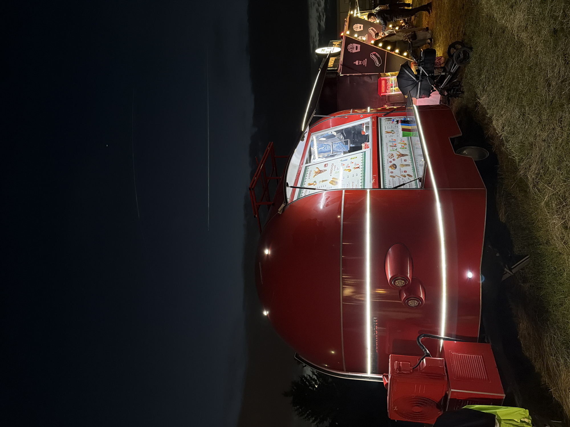 A wide-angle, eye-level shot captures a vibrant red food trailer at dusk. The trailer is illuminated by white LED strip lights that wrap around its curved body and under its service window, casting a warm glow on the surrounding dry grass. A logo with Russian text is visible on the trailer's side. To the right of the main trailer, a smaller, darker food stand is also lit, displaying images of french fries, hot dogs, and drinks. Several people are gathered near the second stand, some appearing to be customers and staff. A black baby stroller is parked in front of the red trailer. The background features a dark sky with faint clouds and at least two visible contrails from aircraft. The scene appears to be set in an outdoor event or market.