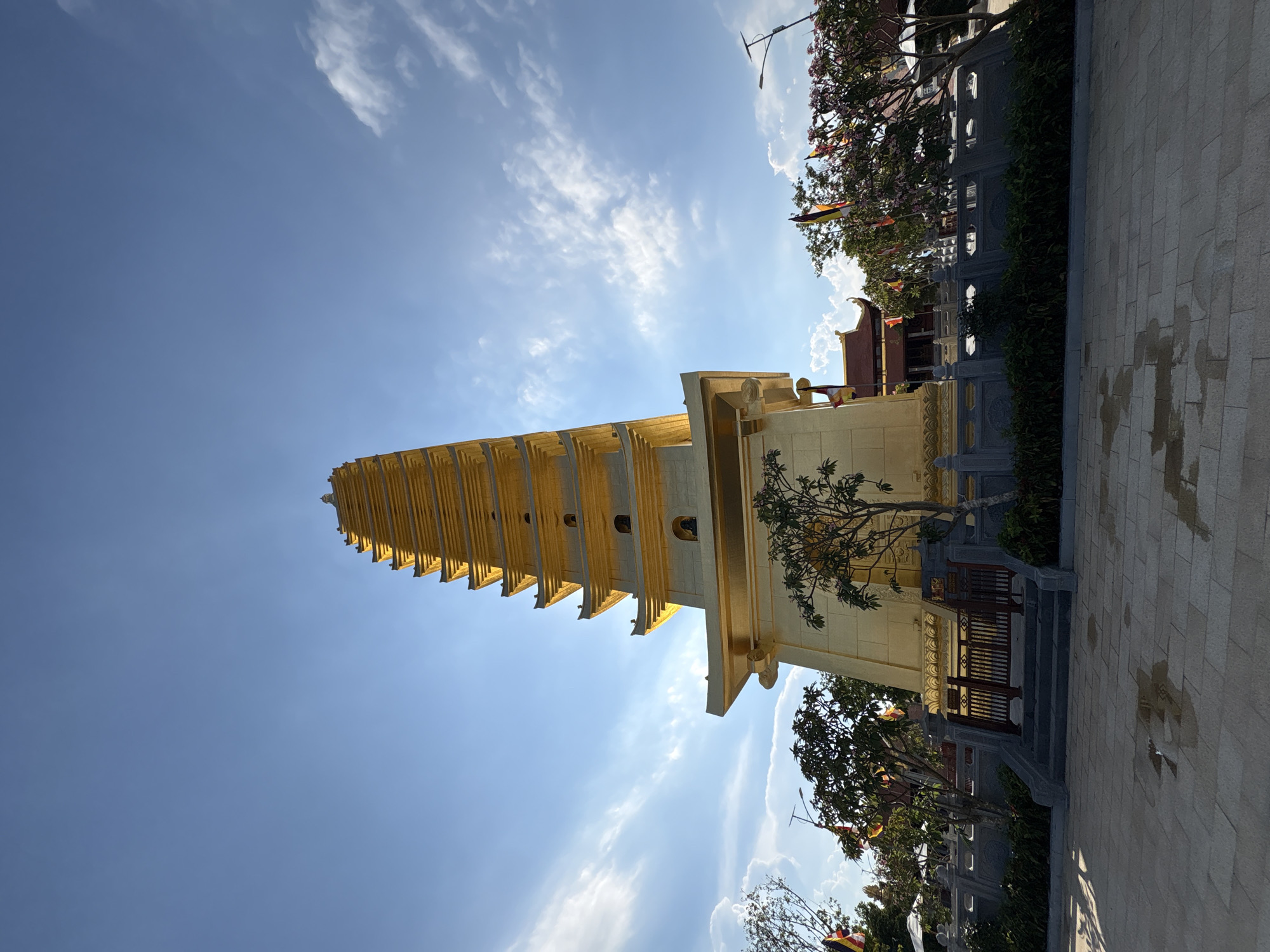 The image captures the Kim Son Pagoda, a prominent golden pagoda located in Diên Khánh, Vietnam. The pagoda's tiered structure reaches into a blue sky dotted with wispy clouds. A small tree grows near the base of the pagoda, next to a wooden railing and steps leading towards the structure. Beyond the pagoda, trees and decorative poles with colorful flags are visible. A paved area is in the foreground.