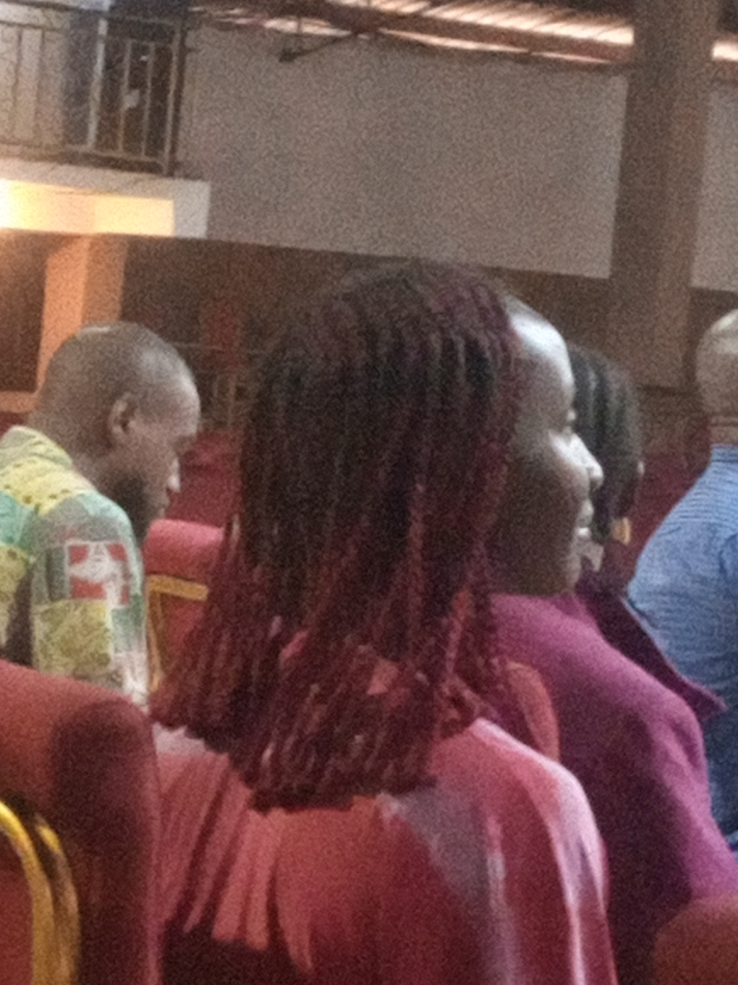A group of people are seated indoors, likely in a place of worship or assembly hall, given the seating arrangement and the overall ambiance. In the foreground, a woman with dark, braided hair is visible from the side, wearing a pink top. To her left, a man with a shaved head and a patterned shirt with green, yellow, and red squares is also seated. Further to the right, another individual wearing a blue shirt can be seen. The background suggests an elevated area with railings and a textured wall, possibly indicating a balcony or upper level. The lighting is dim, with a warm glow suggesting artificial light sources. The scene appears to be captured during a service or event. The location, Rukuba Road, Nigeria, indicates an urban setting.