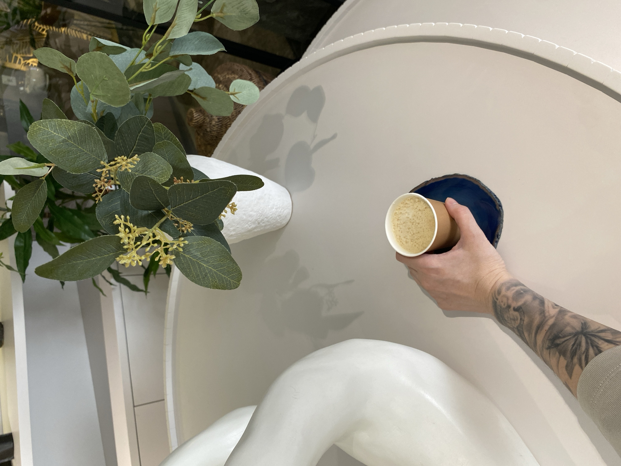 A high-angle shot shows a hand holding a brown paper cup filled with a beige liquid, seemingly coffee, near a circular blue hole in a smooth, cream-colored surface. The hand, with a tattoo covering most of the forearm, appears to be placing the cup into the hole. To the left is a floral arrangement with green leaves and small yellow flowers. The background has a glimpse of dark glass and woven material, suggesting an interior space. There is no information to assume that this image was taken in the city of Votkinsk, Russia.