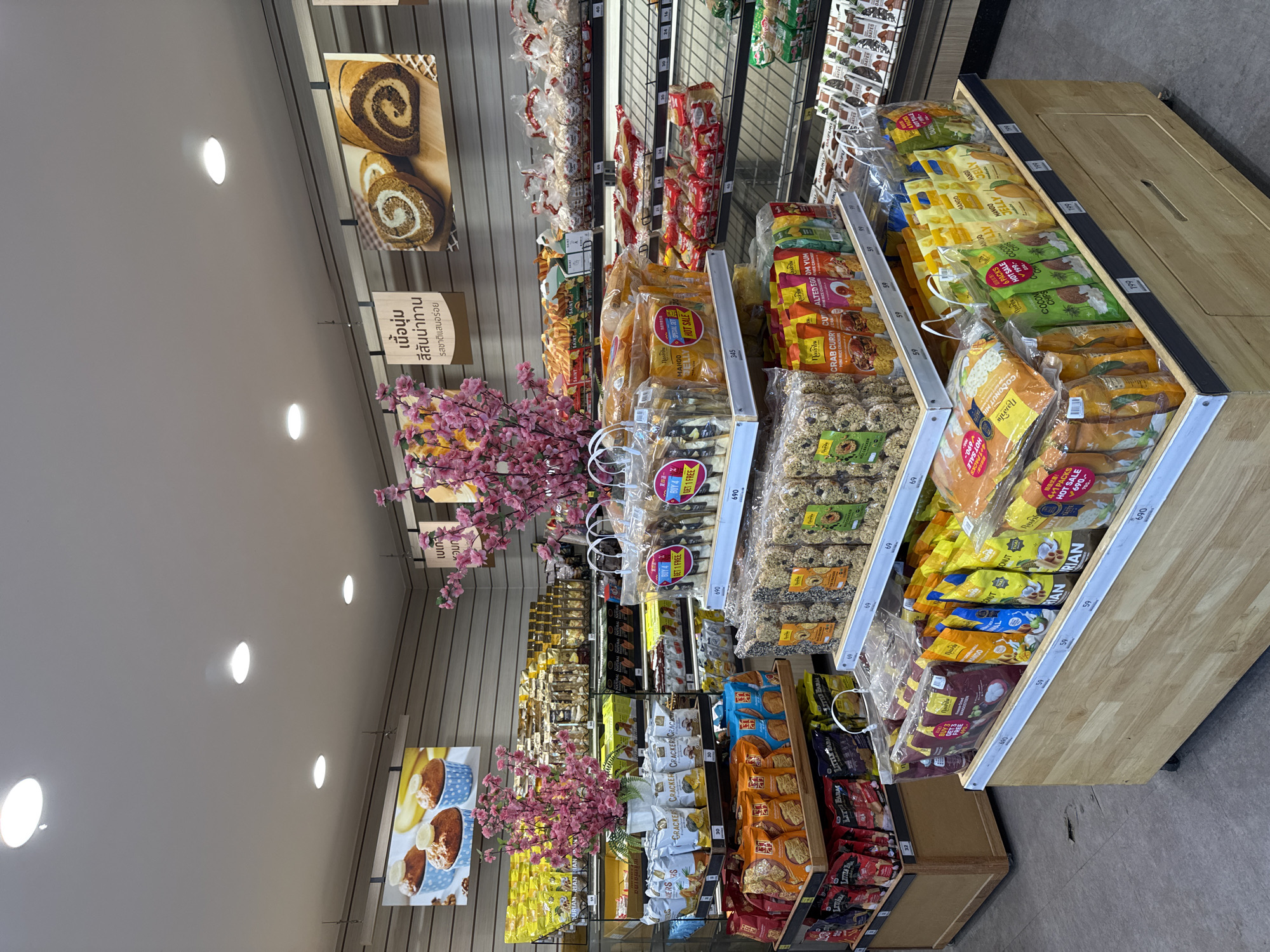 The image captures the interior of a grocery store in Thong Sala, Thailand. The perspective is from the aisle, focusing on shelves stocked with various packaged foods, including cookies, crackers, and jelly. Signage in Thai is visible, indicating product descriptions and promotional offers. Artificial cherry blossom decorations are interspersed throughout the displays, adding a decorative element to the retail environment. Fluorescent lights illuminate the scene from the ceiling.