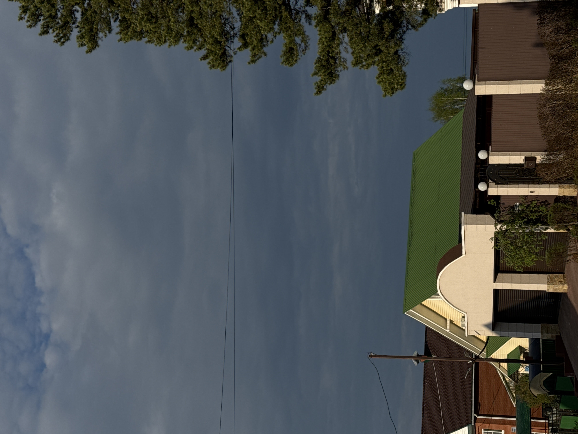 The image shows a view of the sky with clouds, bordered at the top by tree branches. Below, rooftops of houses and buildings are visible. The location is identified as Прикамский, Russia, based on the image's context.