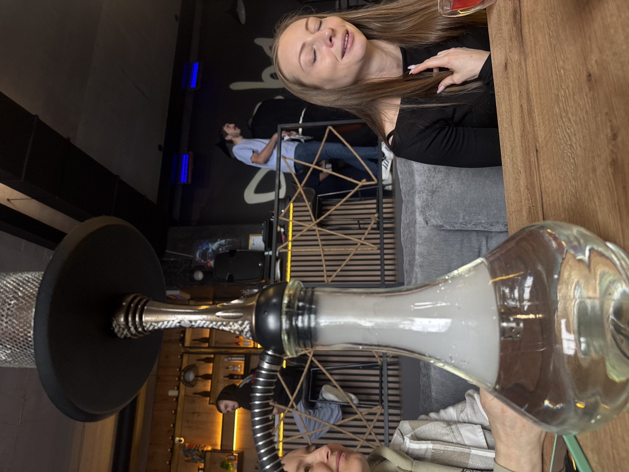 The image captures the interior of a bar or lounge in an unknown city, as indicated by the "Unknown, Unknown" location. Dominating the foreground is a hookah, with its glass base filled with a milky white substance (likely smoke). Two women are partially visible around the hookah. A woman with light brown hair is seen smiling in the upper right. Another person's face is seen in the bottom center.
In the background, a man is standing near a geometrically designed shelf or display. The shelf has decorative rope accents and a bottle or small framed object on its lowest level. Behind the man is a dark wall with light-colored graffiti or design. The lighting in the bar is dim. A segment of a grey sofa is visible near the top right of the frame.