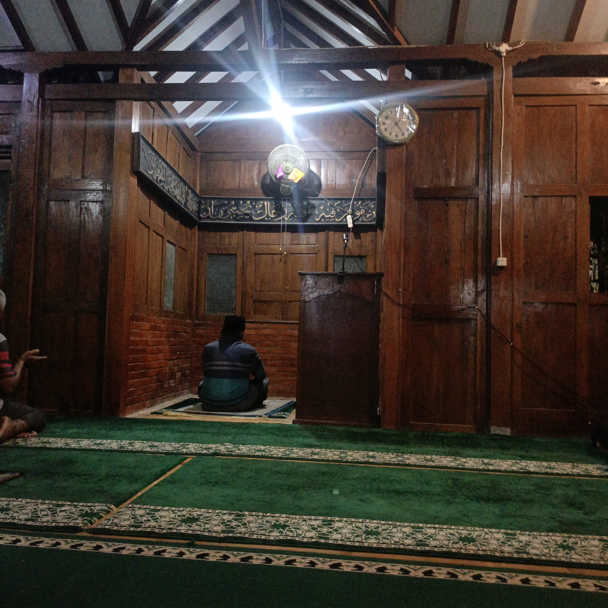 The image shows the interior of a mosque in Tepus, Indonesia. A man wearing a black cap and a striped shirt is seated on a prayer mat on the green carpet. He is facing away from the camera, towards a wooden podium. The mosque has wooden walls and a high, beamed ceiling. A fan is visible on the wall above the man, and a clock is to its right. The overall impression is one of quiet reverence and simplicity.