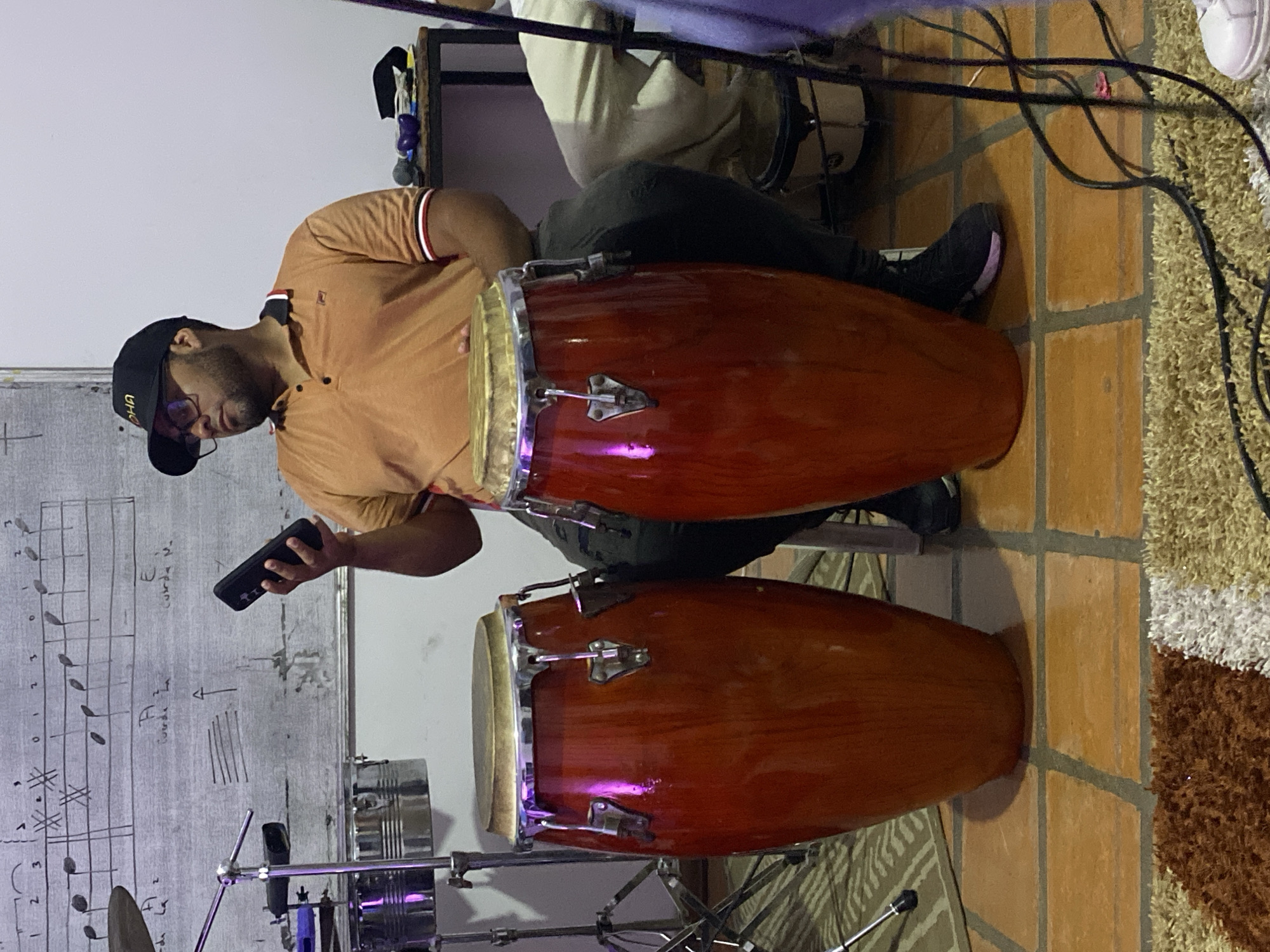 A man wearing a black baseball cap, glasses, and an orange polo shirt is seated behind two conga drums. He is holding a black smartphone in his left hand and appears to be looking at it. The drums are red-brown with chrome hardware and are mounted on a chrome stand. To the left of the man is a white board with musical notes and annotations. Behind the man, another person is partially visible seated behind a drum, and there are cables and other musical equipment. The floor is covered with brown and beige tiles and a patterned rug. The location is Tinaquillo, Venezuela.