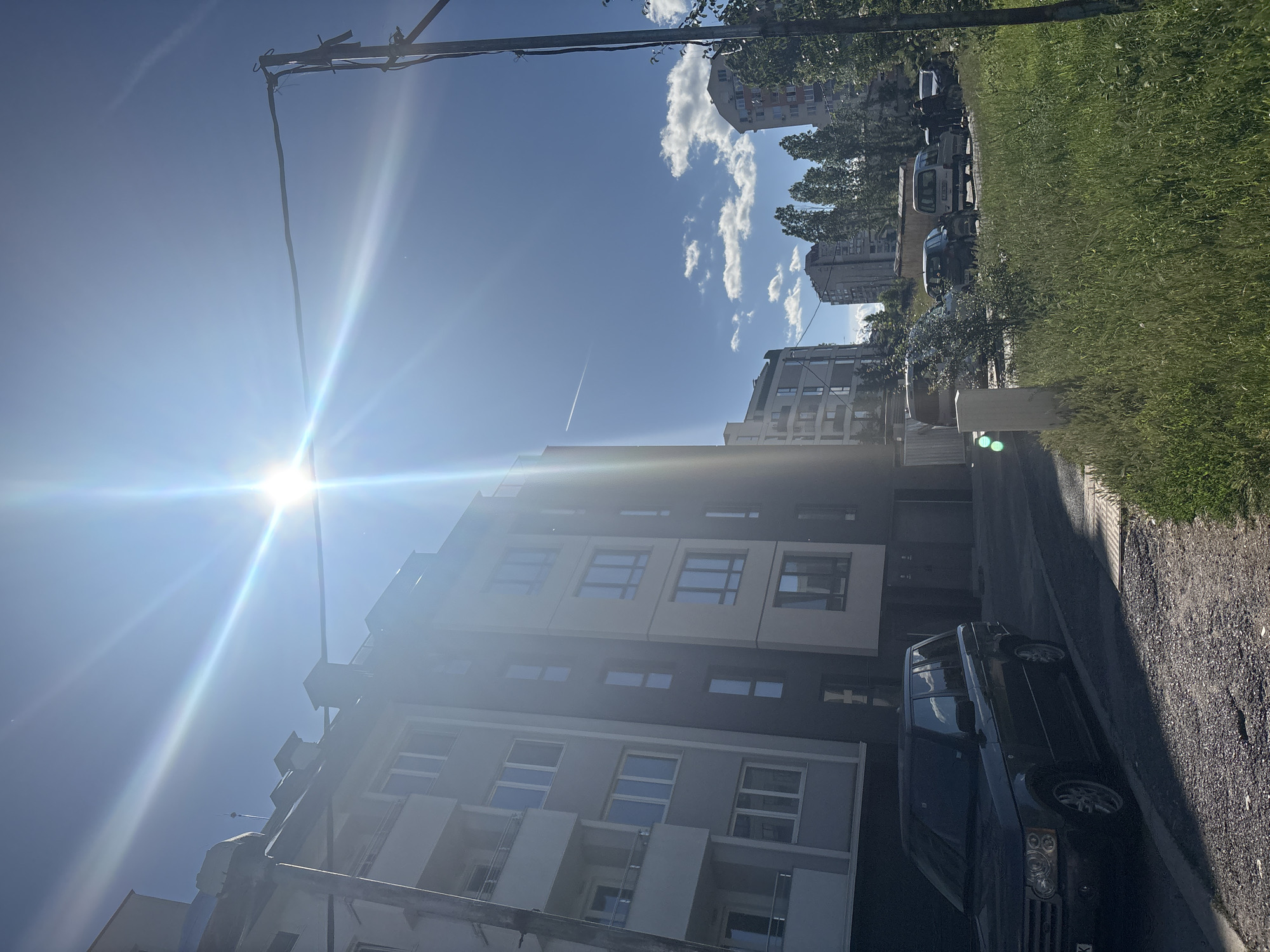 A wide-angle shot captures a sunny urban scene with modern apartment buildings under a bright blue sky. The sun is high and flares brightly, creating lens flares that extend across the upper portion of the image. In the foreground, a dark-colored SUV is parked on the side of a paved road. Behind the SUV, a multi-story apartment building with a light gray and darker gray facade dominates the left side of the frame. To the right of this building, another apartment building with a beige and dark gray facade is visible. Further in the background, several other buildings of varying heights and architectural styles, interspersed with trees, are present. A thin, white contrail from an airplane is visible in the sky. Several cars are parked in a row along the street in the mid-ground. Green grass and some foliage are present on the right side of the image, growing along the side of the road. A thin, dark electrical wire is suspended horizontally across the upper right portion of the image, supported by poles.