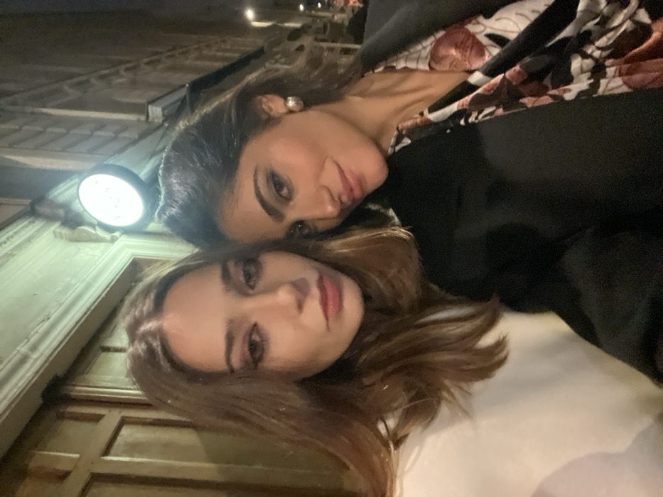 Two young women pose closely together. One has dark hair, a black blazer, and a floral top. The other woman has lighter brown hair. They are illuminated by a bright street light mounted on a building façade with ornate trim. The scene is taken from a low angle, giving a tilted perspective of the women and the building.