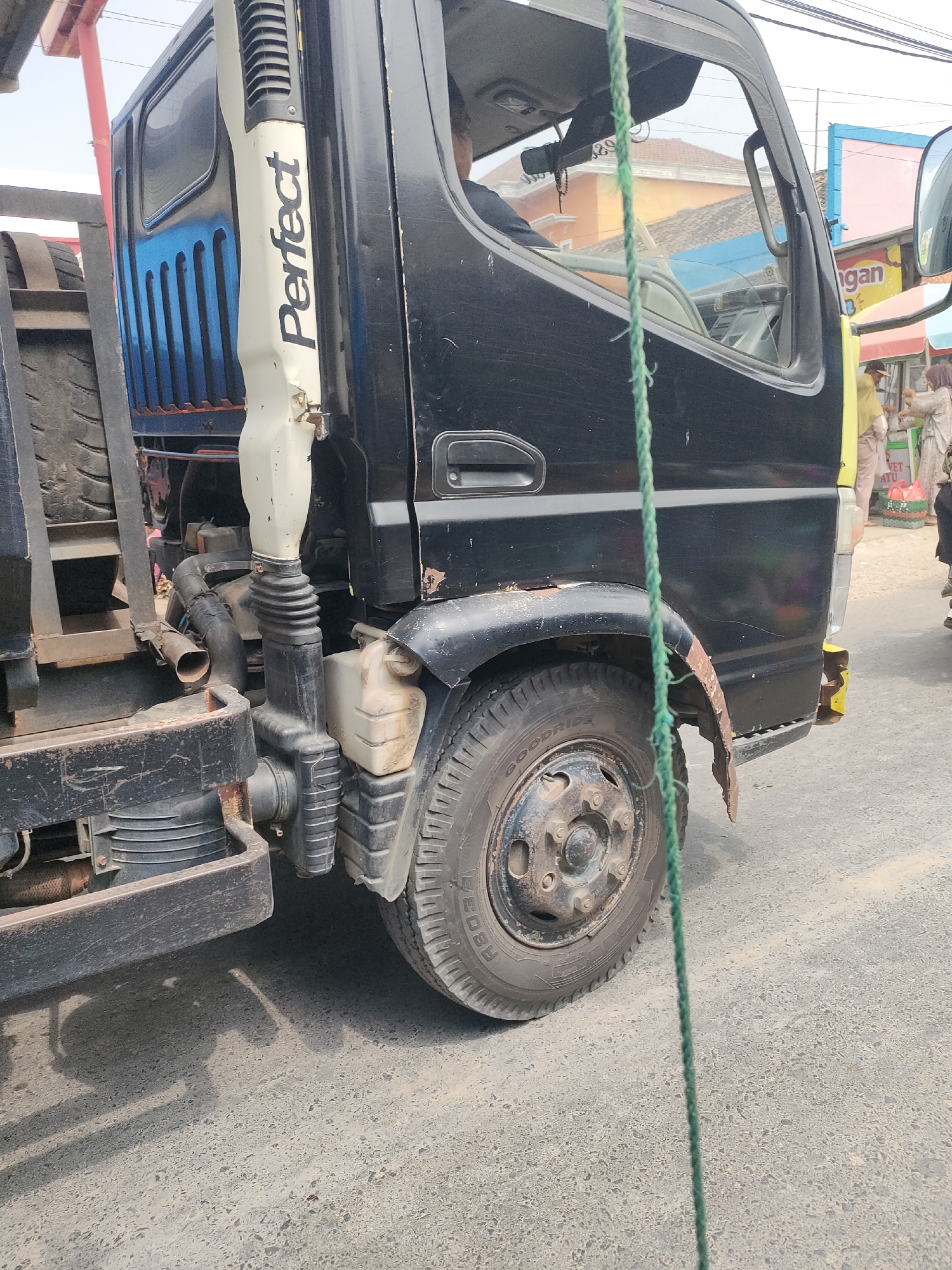 The image shows a close-up, low-angle view of a black truck parked on a paved road in Sukoyoso, Indonesia. The truck's front passenger-side wheel and fender are prominent in the foreground. A white vertical piece with the word "Perfect" in black lettering is attached to the truck's body. The truck exhibits signs of wear, with rust visible on the fender and yellow paint on the bumper. A green rope hangs vertically in the center of the frame, obscuring parts of the truck. In the background, buildings and a glimpse of people in a market area are visible. The overall environment appears to be a bustling street scene.
