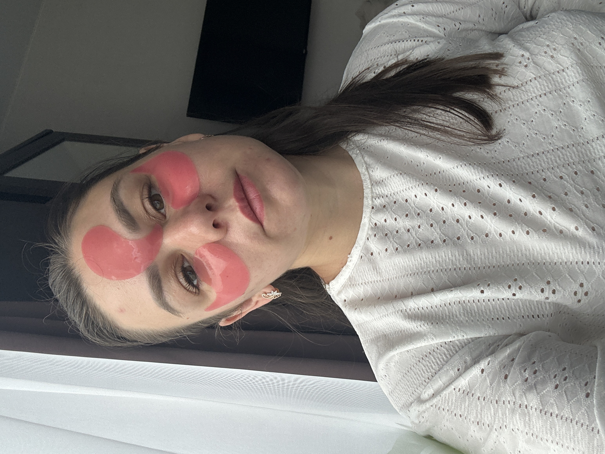 A young woman with brown hair is taking a selfie in a well-lit indoor setting, possibly her home, located in Chaikovsky, Russia. She is wearing a white, intricately patterned long-sleeved shirt and has pink under-eye patches applied to her face, with one patch extending slightly onto her forehead, creating a unique decorative effect. The woman's expression is neutral, and she is looking directly at the camera. Her hair is pulled back, with some strands falling loose around her face and shoulders. The background is softly blurred, with a white curtain visible on the left and a dark television screen or similar object on the right. The overall ambiance is casual and relaxed, suggesting a moment of personal care or a break in her day.