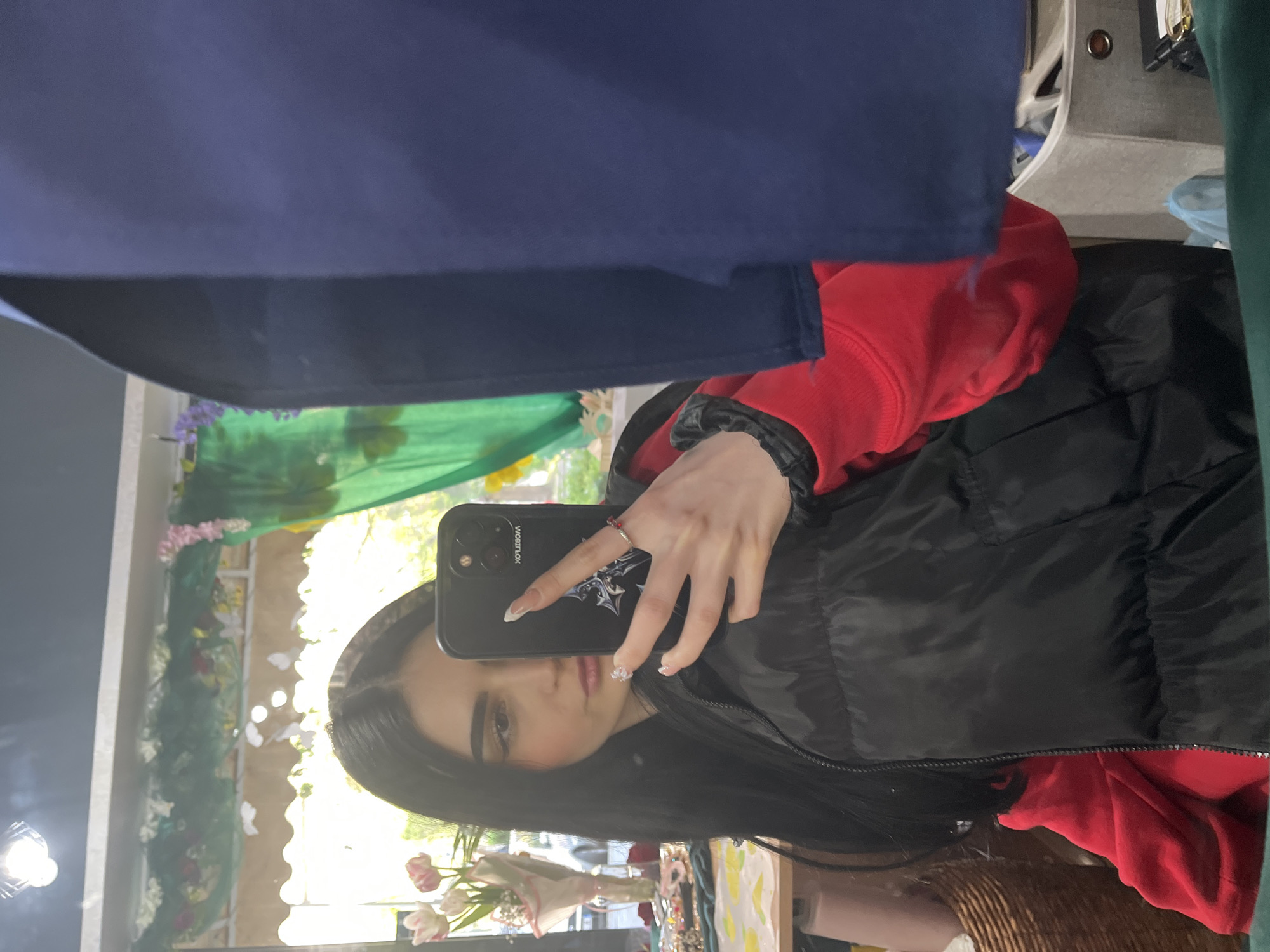 A young woman with dark hair takes a selfie in a mirror. She holds a black smartphone in her right hand, partially obscuring her face. She wears a red hooded sweatshirt under a black puffer jacket. The reflection in the mirror shows a decorated window with floral arrangements, possibly indicating a shop or indoor market setting. Based on the image content alone, there is no verifiable information to pinpoint the exact location within Burgas, Bulgaria.