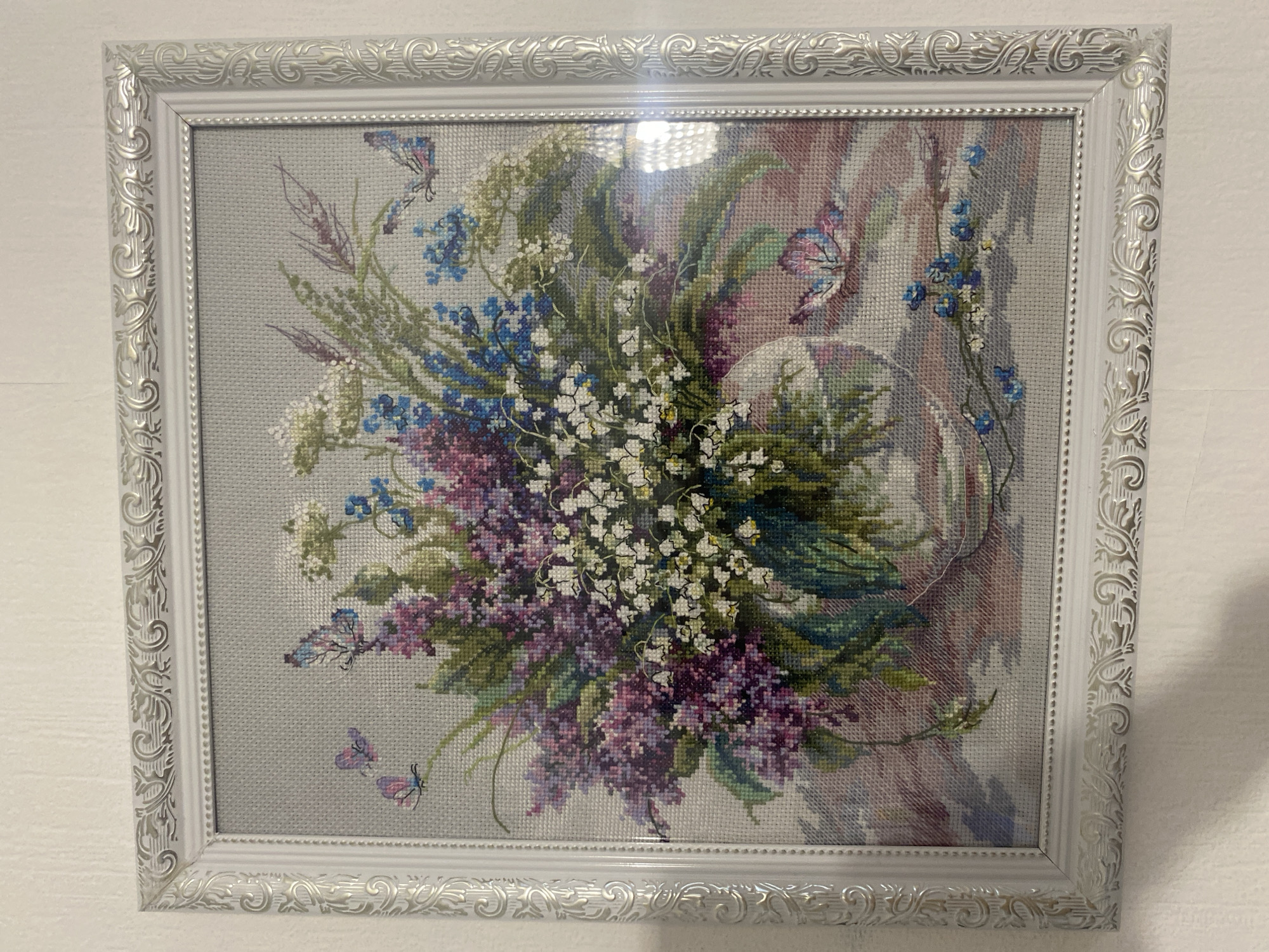 The image shows a framed cross-stitch embroidery of a floral arrangement featuring white, blue, and purple flowers, along with green foliage and butterflies, set against a gray background. The embroidery is within a white decorative frame with ornate detailing. Light reflection obscures a small area in the center. The background appears to be a plain, white wall. There is no indication of a specific location in Yekaterinburg, Russia, or any individuals present in the image.