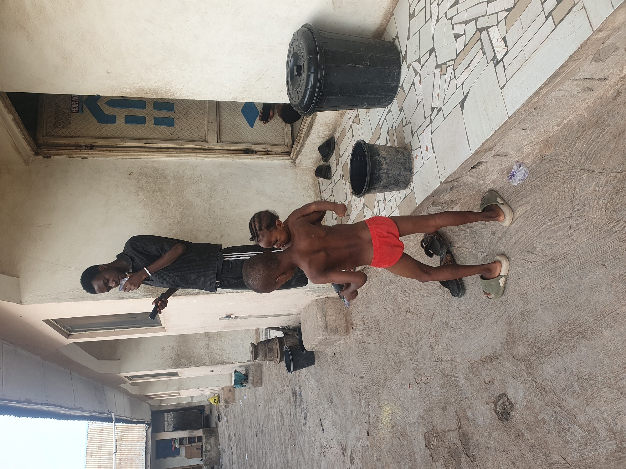 This image depicts an outdoor or semi-outdoor area, likely in a residential or communal setting in Lokoja, Nigeria, under bright daylight conditions. The ground is a mix of rough concrete and an area paved with a mosaic pattern of broken white and grey tiles.
Three individuals are present: an adult male and two young children. The adult, dressed in a dark striped shirt and dark pants, stands on the left, engaged with a smartphone or similar device held in his hand. He wears a white bracelet on his right wrist. To his right are two children. The taller child, appearing to be a boy, is shirtless and wears red shorts. He stands facing and looking towards the smaller child. The smaller child, a girl with braided hair, is also shirtless and positioned in front of the boy, seemingly interacting with him. Various sandals and slippers are scattered on the concrete ground, including a pair of green slides and dark slides.
In the background, against a light-colored wall, stand two black plastic buckets—one with its lid on and the other open. A small blue cloth or sponge is visible near the open bucket. Further back, a dark recessed area features a metal surface, possibly a shuttered window or door. On this metal surface, a blue logo is visible, comprising an arrow pointing left and several rectangular shapes. Partially visible text next to this logo reads "FEAR NO". The scene captures a casual, everyday moment with no strong discernible emotions displayed by the subjects.