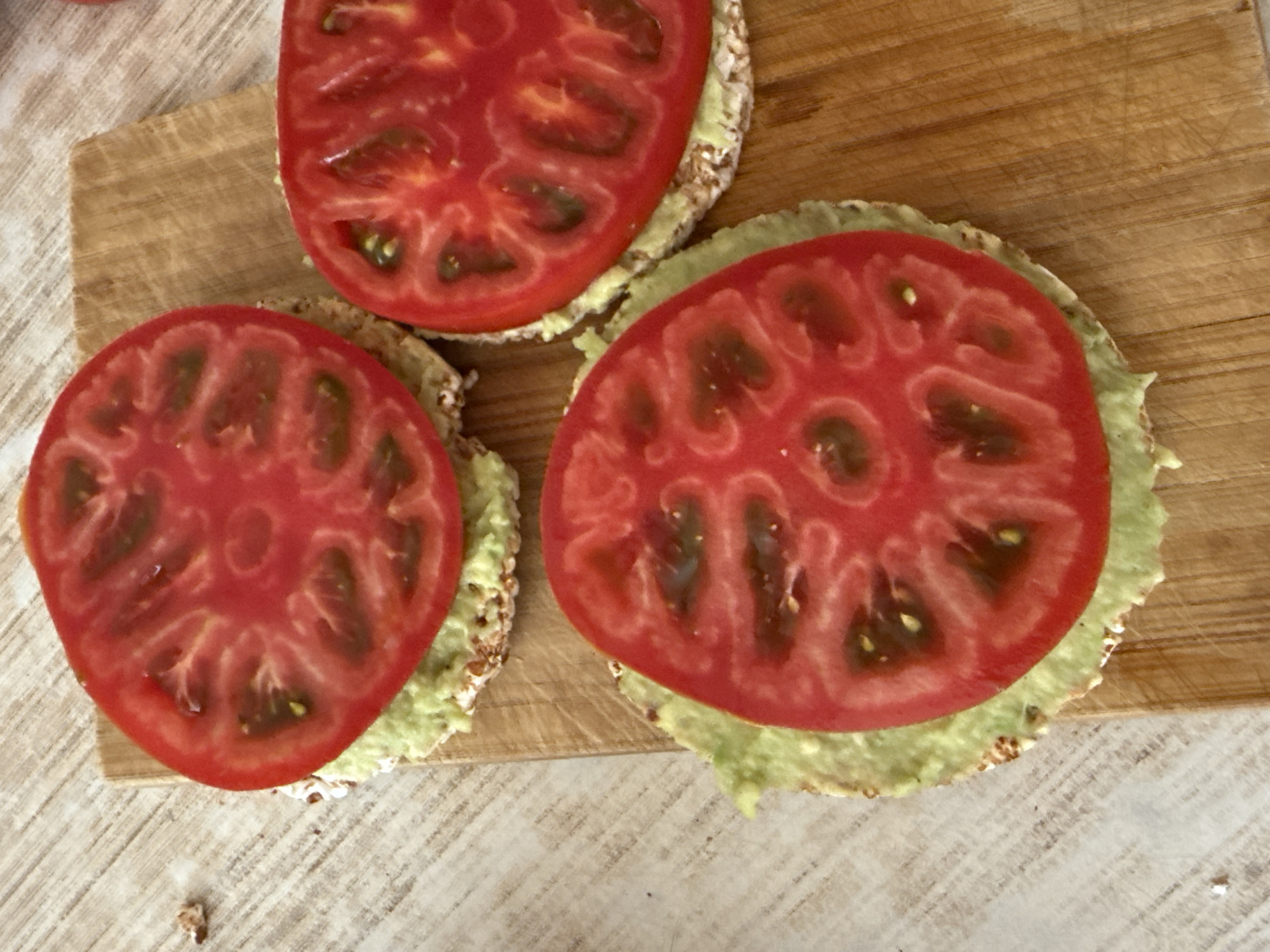 This is a close-up, overhead shot of three open-faced snacks on a wooden cutting board. Each snack consists of a round, puffed grain cracker topped with a light green spread, likely mashed avocado, and a thick slice of ripe red tomato. The tomatoes are cut crosswise, revealing their juicy interiors and seeds. The top snack is partially out of frame, while the other two are fully visible. The wooden cutting board has visible grain and scratch marks, suggesting it is well-used. The background surface is a light-colored, textured material, possibly a tablecloth or countertop. The lighting appears to be natural, casting soft shadows. The overall impression is one of a simple, healthy snack being prepared or served. No people are visible, and there is no visible text. The scene suggests a casual meal or snack time.