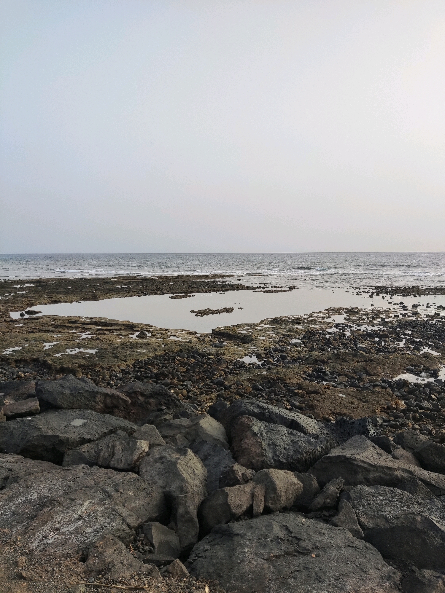 The image depicts a rocky coastline with the ocean in the background. The foreground is dominated by large, dark grey rocks. In the mid-ground, there is a flatter, more varied rocky area with shallow pools of water. The ocean stretches to the horizon under a pale, overcast sky. The scene appears to be a natural coastal landscape. While the prompt suggests the location is Arona, Spain, there are no specific visual cues in the image to confirm this. There are no people, animals, or man-made structures visible.