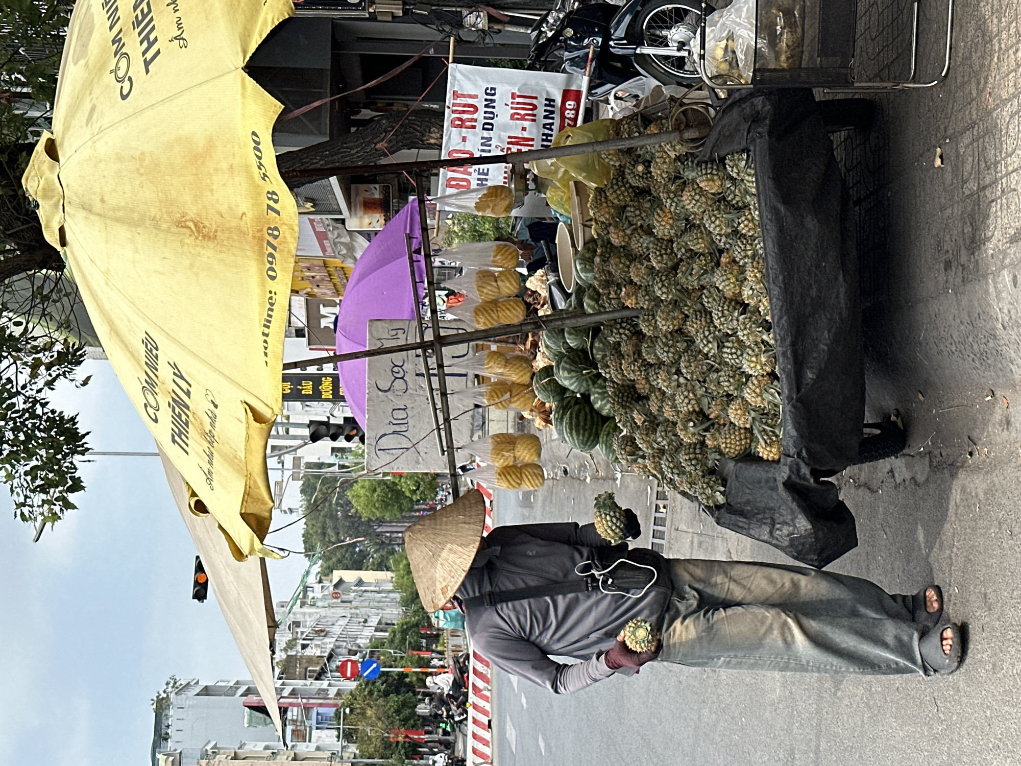 Here is a detailed description of the image:
A street vendor in Ho Chi Minh City, Vietnam, stands beside his pineapple cart. The cart is laden with many pineapples and has a yellow umbrella attached to provide shade. The vendor wears a traditional conical hat, a gray long-sleeved shirt, long pants, and sandals. He is holding a pineapple in each hand. A sign on the cart advertises "Dao-Rut" and includes a phone number. A purple umbrella is also visible. In the background, city buildings, traffic lights, and motorbikes are seen on the street.