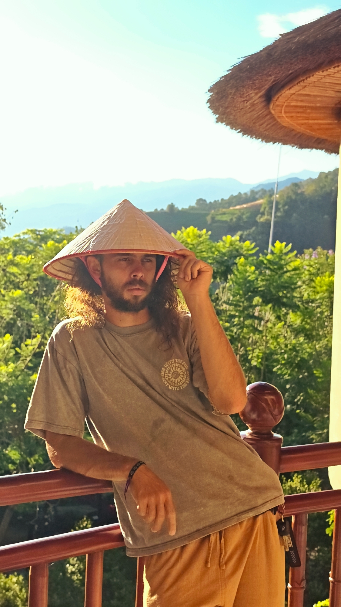 A man in his 20s or 30s is captured in a sunlit outdoor setting, leaning against a red wooden railing. He has long, dark, curly hair and a beard, and wears a traditional Vietnamese conical hat (nón lá) with a pink chin strap, which he is holding with his right hand. His gaze is directed thoughtfully towards the left. He is dressed in a faded greyish-green t-shirt featuring a circular graphic on the left chest area, which includes text like "RISE WITH THE SUN" and "SHINE WITH THE SUN" surrounding a sun or mandala-like design. He also wears mustard yellow or tan drawstring shorts, and a dark bracelet on his left wrist. A black lanyard or keychain with the text "GCS" and downward-pointing arrows is visible hanging from his shorts.
The scene is bathed in warm, bright sunlight, suggesting either early morning or late afternoon. The background is lush with vibrant green foliage, appearing as hills or cultivated terraces, and distant mountains under a clear, bright blue sky with some atmospheric haze. Part of a thatched roof structure is visible in the upper right corner, contributing to the rustic, traditional ambiance. The overall impression is one of serene observation amidst the scenic landscape of Mu Cang Chai, Vietnam.