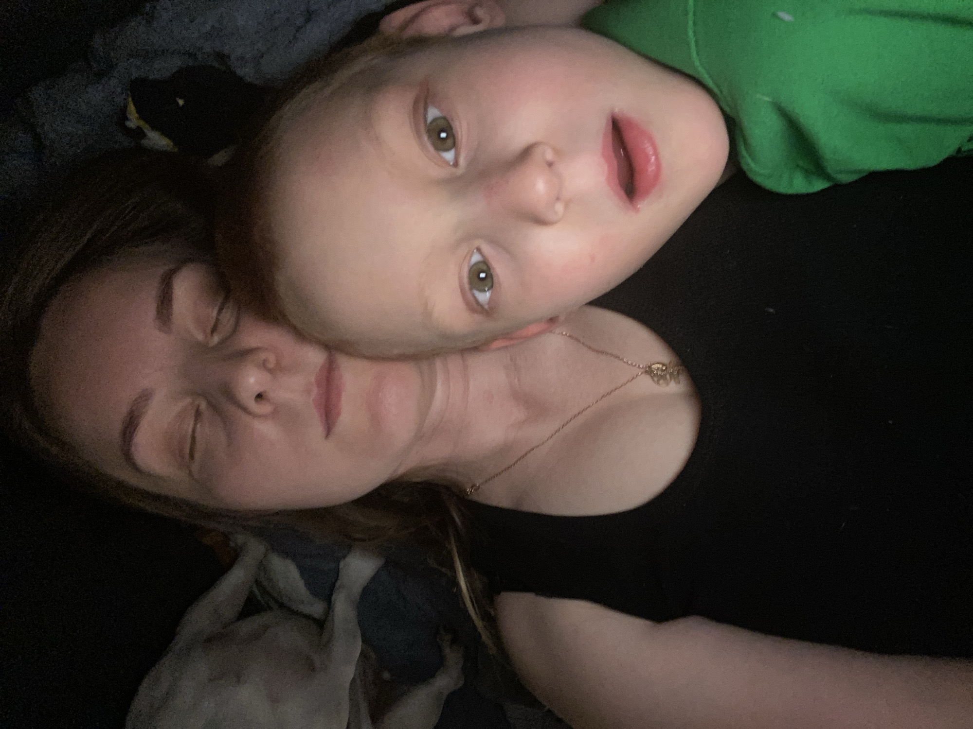 A woman with closed eyes lies on a dark surface, presumably a bed, next to a child with open eyes. The child is wearing a green shirt. The woman is wearing a black shirt and a necklace. The background appears dark and cluttered. A black and white object is visible in the top left corner. There is no indication of location in Noviy, Russia.
