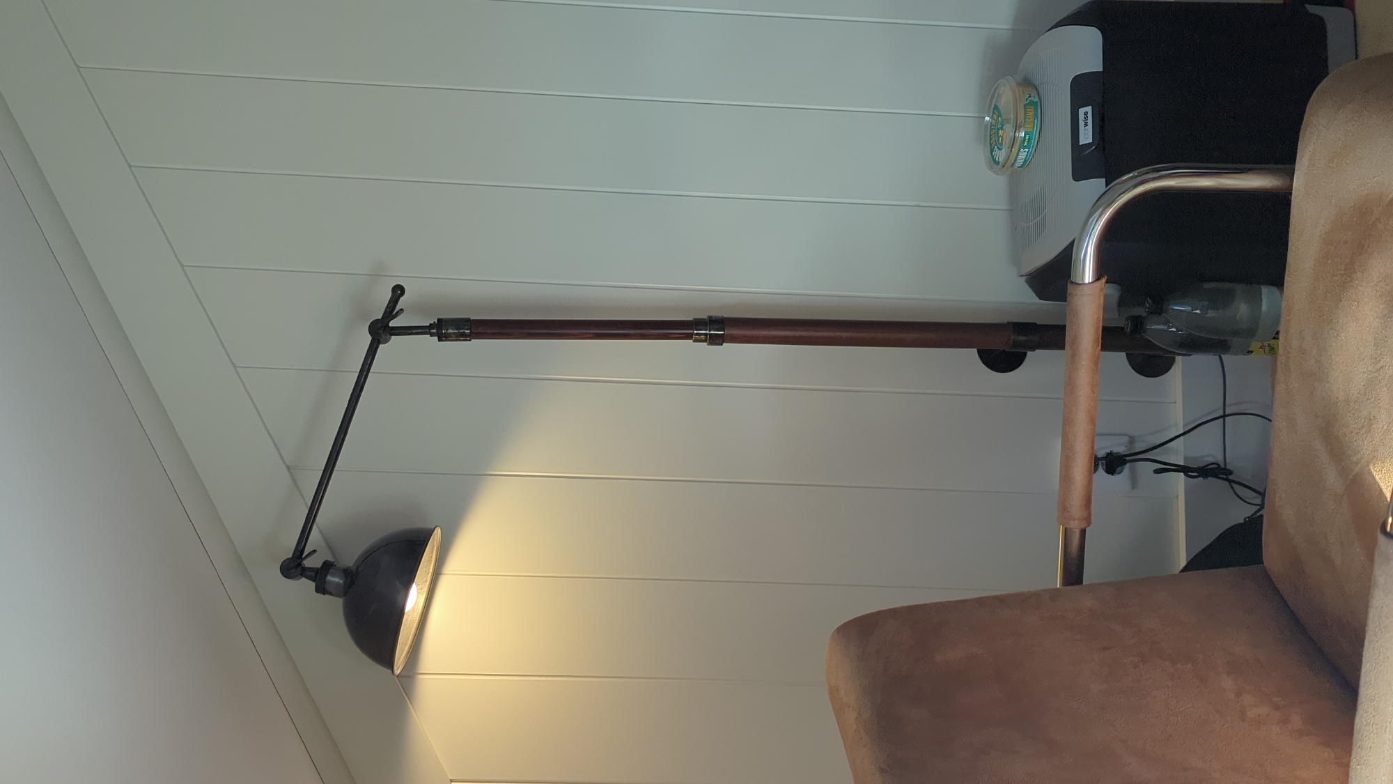 The image shows an indoor scene with a floor lamp, a chair, and a small cooler. The floor lamp is dark metal with a single dome-shaped shade emitting a warm light. It has an adjustable arm and a tall, slender pole that appears to be made of dark wood with metal accents. The chair in the foreground is upholstered in a light brown, textured fabric, with chrome-plated armrests. To the right of the lamp and chair, a small black and grey cooler is visible. On top of the cooler sits a round, clear plastic container with a label that appears to be for food, possibly a spread or dip. The background consists of a white wall with vertical paneling. There are two plastic bottles near the base of the floor lamp. The scene is indoors, and given the presence of a cooler and the general interior design, it could be a room in a house or a cabin. The location is specified as Larkollen, Norway.