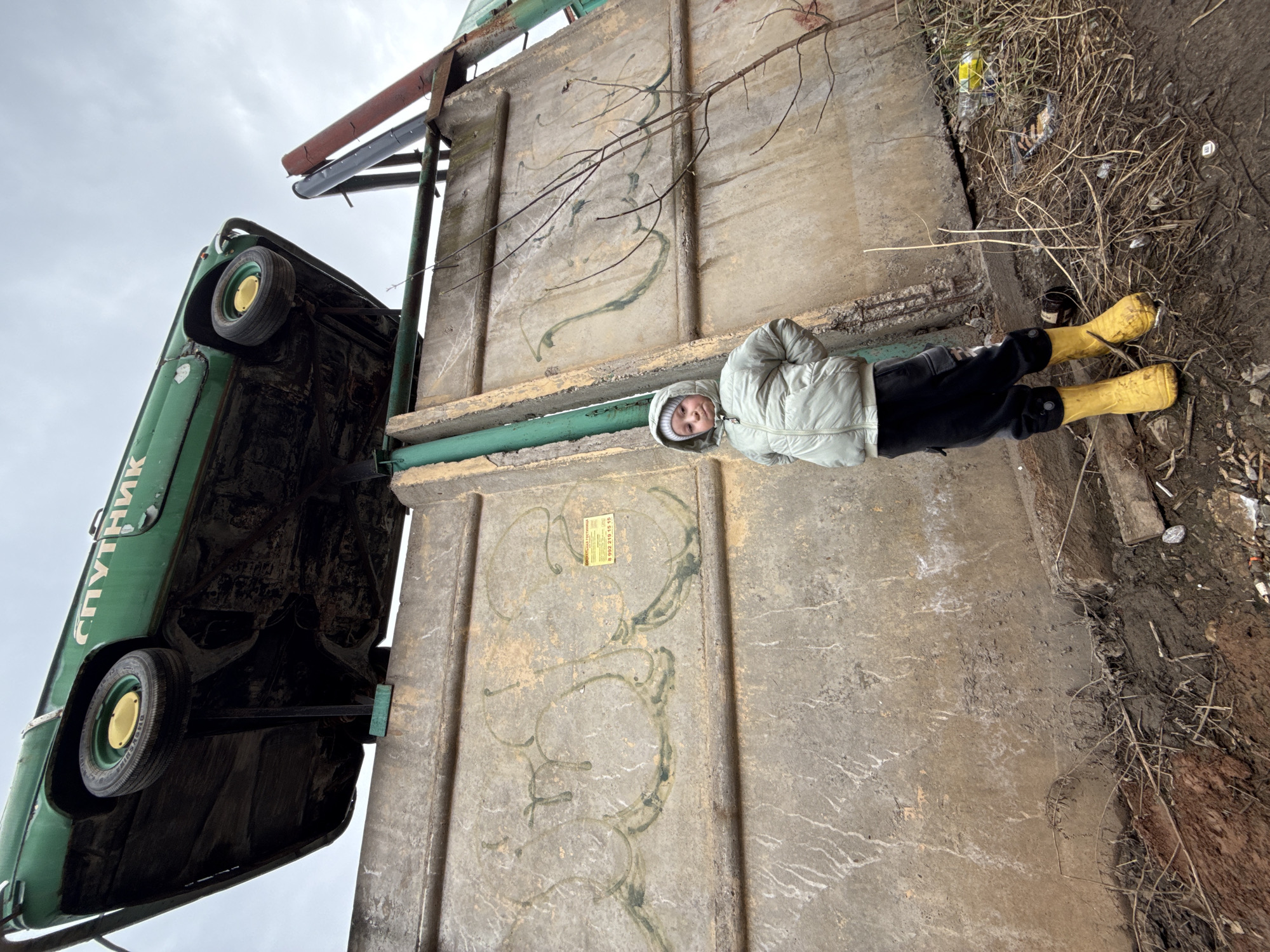 A child in a hooded jacket and yellow boots stands next to a concrete structure that suspends a green car in the air. The car has the word "СПУТНИК" (Sputnik) written on it in Cyrillic letters. The ground around the child is dirt and debris. The concrete structure has graffiti on it. The sky is overcast. The location is likely Perm, Russia, given the Sputnik reference and Cyrillic writing.