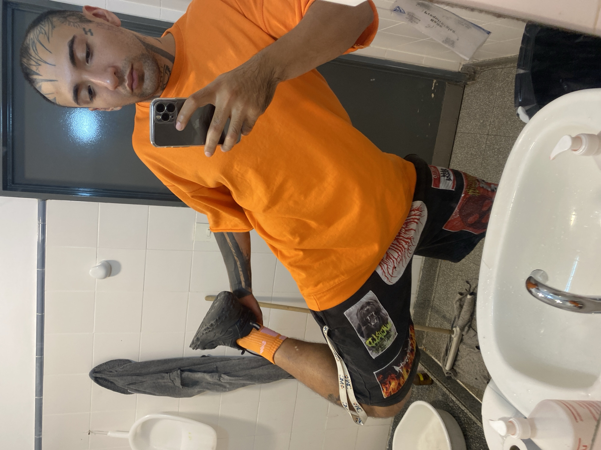 A person with tattoos on their face, neck, and arms is taking a selfie in a bathroom. They are wearing an orange t-shirt, black shorts with patches, and orange socks with grey sneakers. The bathroom has white tile walls, a grey door, a white sink, and a white urinal. A grey cloth is draped near the urinal. The location appears to be a public restroom. There is no evidence in the image to suggest this is in the city of Buenos Aires, Argentina.