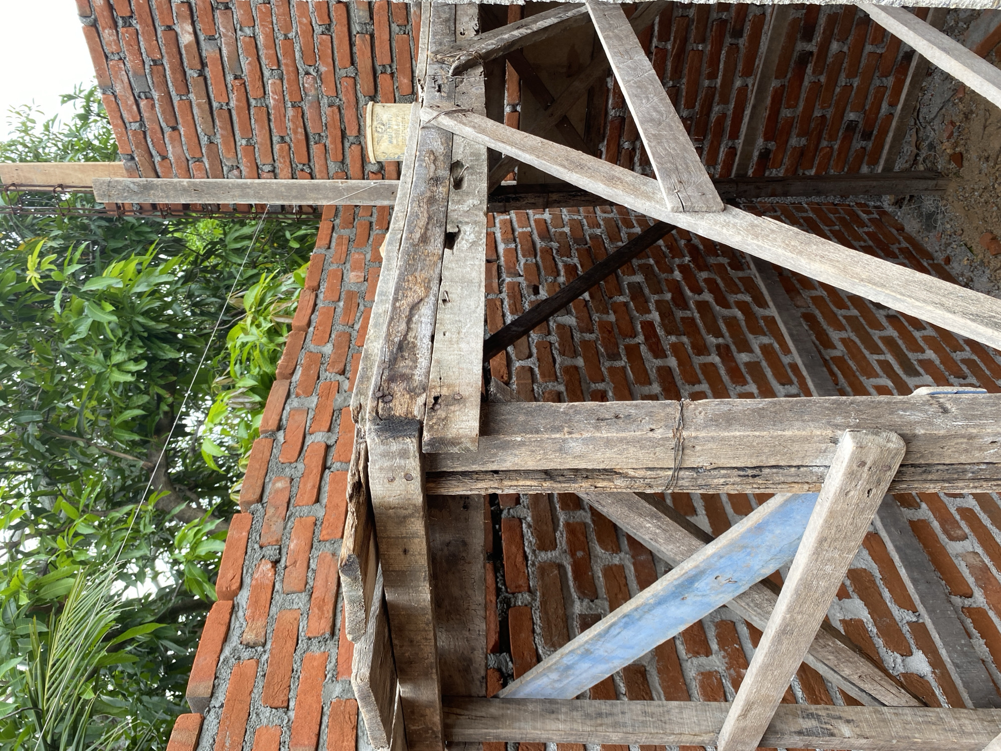 The image shows a construction site in Banda Aceh, Indonesia, focusing on scaffolding and brickwork. The scene depicts a section of a building under construction, with walls made of red bricks and mortar. Wooden scaffolding, constructed from rough-hewn planks and beams, supports a platform overhead. Some of the wooden elements show signs of wear and tear, with nails and wire visible. A pale yellow bucket is present on the platform. Lush green trees and foliage are visible in the background, suggesting an outdoor environment. The ground beneath the scaffolding appears to be covered in dirt and small stones. There are no people visible in the image.