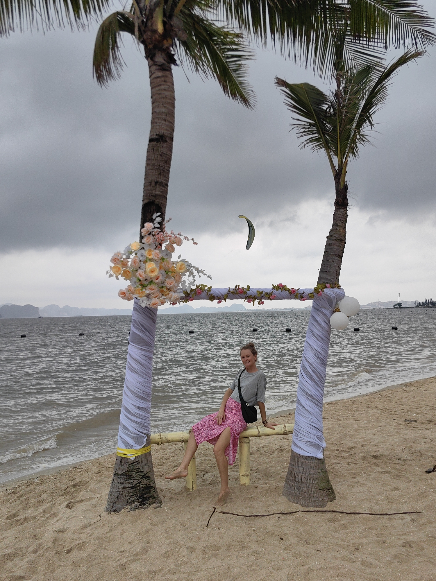 This media file captures a coastal scene in Bai Chay Ward, Vietnam, on an overcast day. In the foreground, two prominent palm trees stand on a sandy beach. Their lower trunks are decoratively wrapped in white, sheer fabric, with a yellow tie visible on the left tree. A light-colored bamboo bench or swing is suspended between the two palms, and a woman is seated barefoot upon it. She has short, light-colored hair and is wearing a grey t-shirt, a pink polka-dotted skirt, and a black shoulder bag, looking towards the viewer with a slight smile.
The palm trees are adorned with floral decorations, suggesting a special event. The left tree features a large cluster of light orange, pink, and white roses and other blossoms. The right tree has a garland of pink roses and green leaves draped across the bamboo structure, and two white balloons are tied to its trunk.
Beyond the beach, the choppy, grey-blue sea stretches towards a horizon line dotted with distant islands or mountains, characteristic of the region's karst topography. Several dark buoys are visible in the water. High in the cloudy sky, a green and grey kite, likely used for kitesurfing, is noticeable above the right palm tree. The weather appears overcast, indicated by the uniformly grey sky and subdued lighting. There is no visible text in the image.