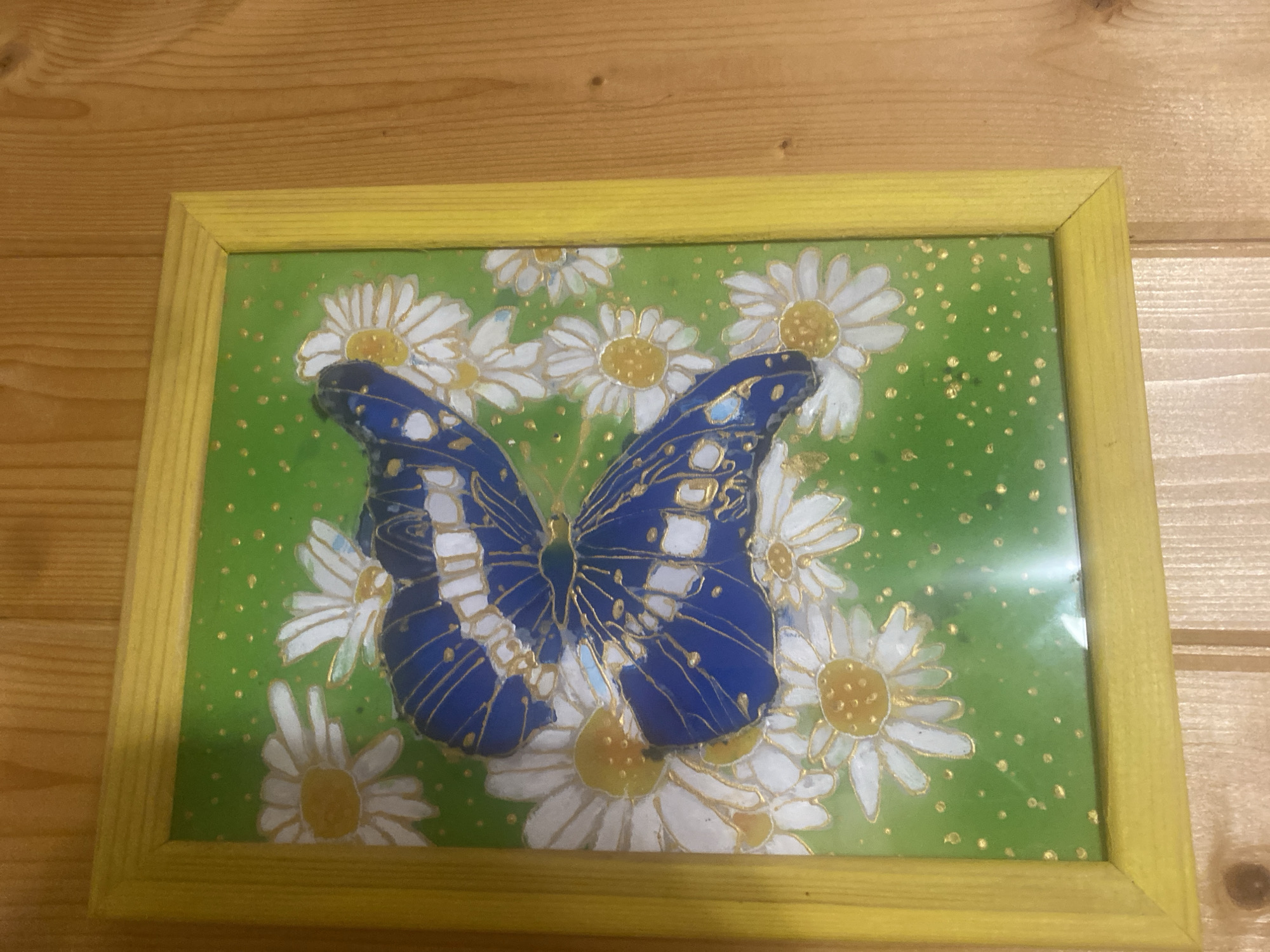 A framed painting of a blue butterfly surrounded by daisies sits on a wooden surface. The painting has a bright green background with gold flecks. The butterfly's wings are predominantly blue with white markings and outlined in gold. The daisies are white with yellow centers, also outlined in gold. The frame is a bright yellow. The wooden surface appears to be a tabletop or shelf.