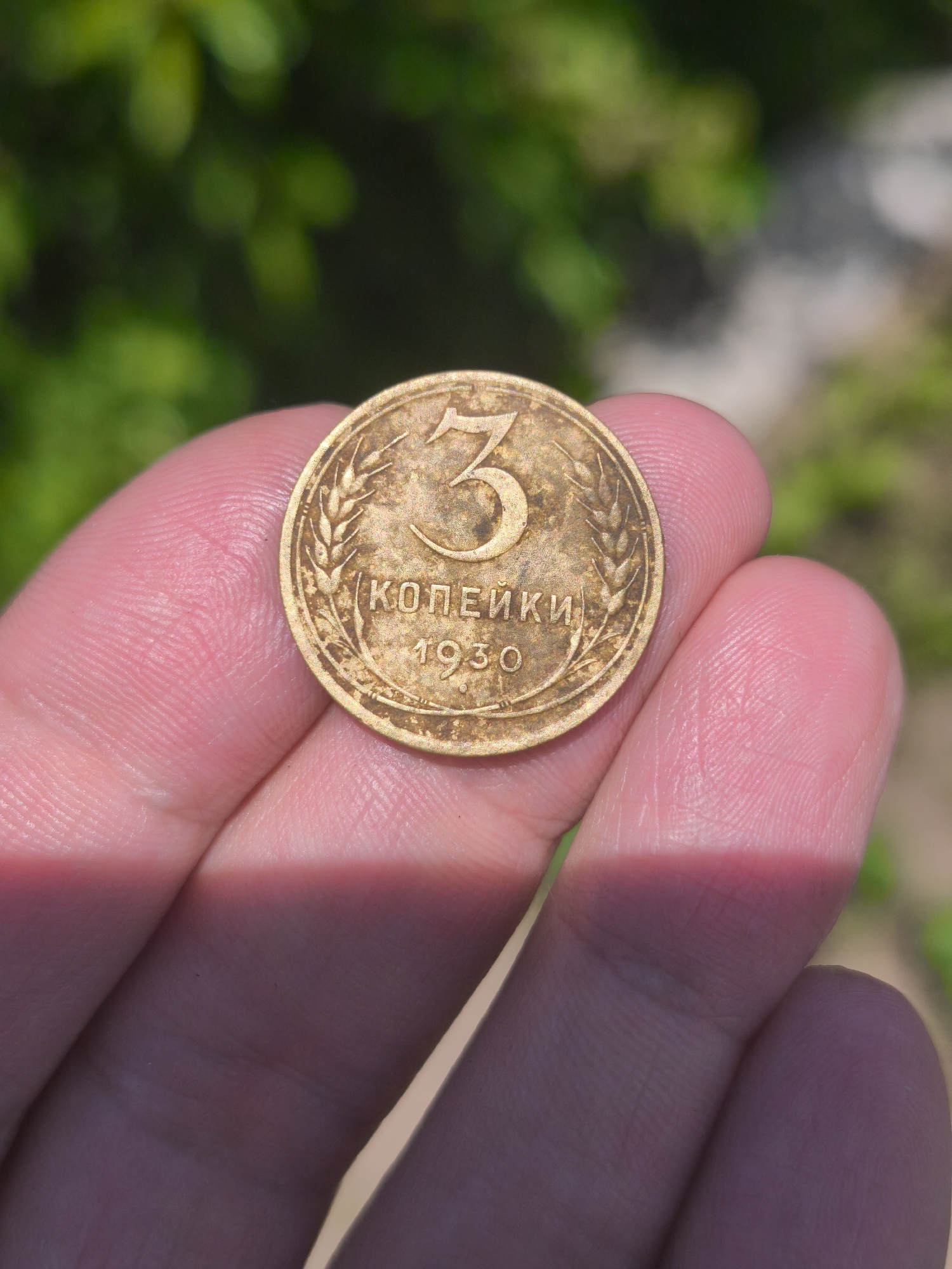A close-up, outdoor, overhead shot shows a human hand holding a weathered, bronze-colored coin. The coin is round and has embossed markings. The number "3" is prominently displayed in the upper center, followed by the Cyrillic text "КОПЕЙКИ" below it, and the year "1930" at the bottom. The coin is surrounded by a decorative border, possibly wheat stalks. The hand holding the coin has visible fingerprints and is illuminated by sunlight casting a bright shadow across the fingers. The background is softly blurred, showing green foliage, suggesting an outdoor setting, possibly in a garden or park. There is no indication of the city of Kady, Russia, in the image.