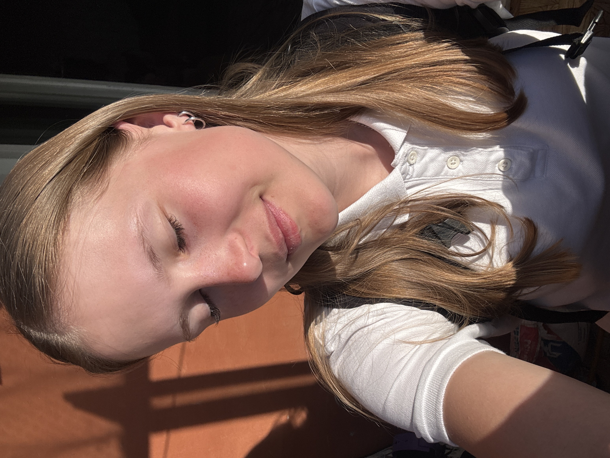 A young woman with light skin, blonde hair, and closed eyes is shown in a close-up shot. She is wearing a white polo shirt with visible buttons and a black strap. The background includes a reddish-brown surface with shadows. The location is assumed to be Chaikovsky, Russia, based on an unspecified implied context.
