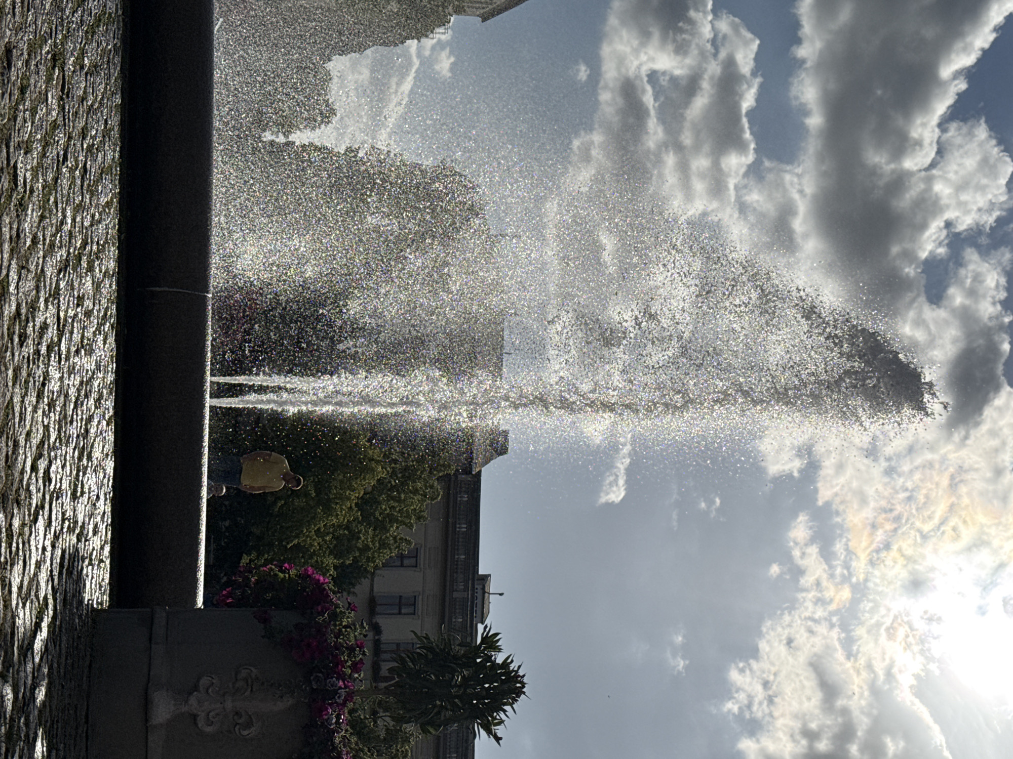 The image depicts a large fountain in a city square, with water spraying high into the air, catching the sunlight. In the background, there are buildings and trees. A person wearing a yellow shirt is standing near the fountain, and a small child is visible behind them. The ground is paved with cobblestones, and a large planter filled with purple flowers is in the foreground on the right. The sky is cloudy, with the sun shining through the clouds. This scene is located in Potsdam, Germany, specifically at the Brandenburger Tor fountain.