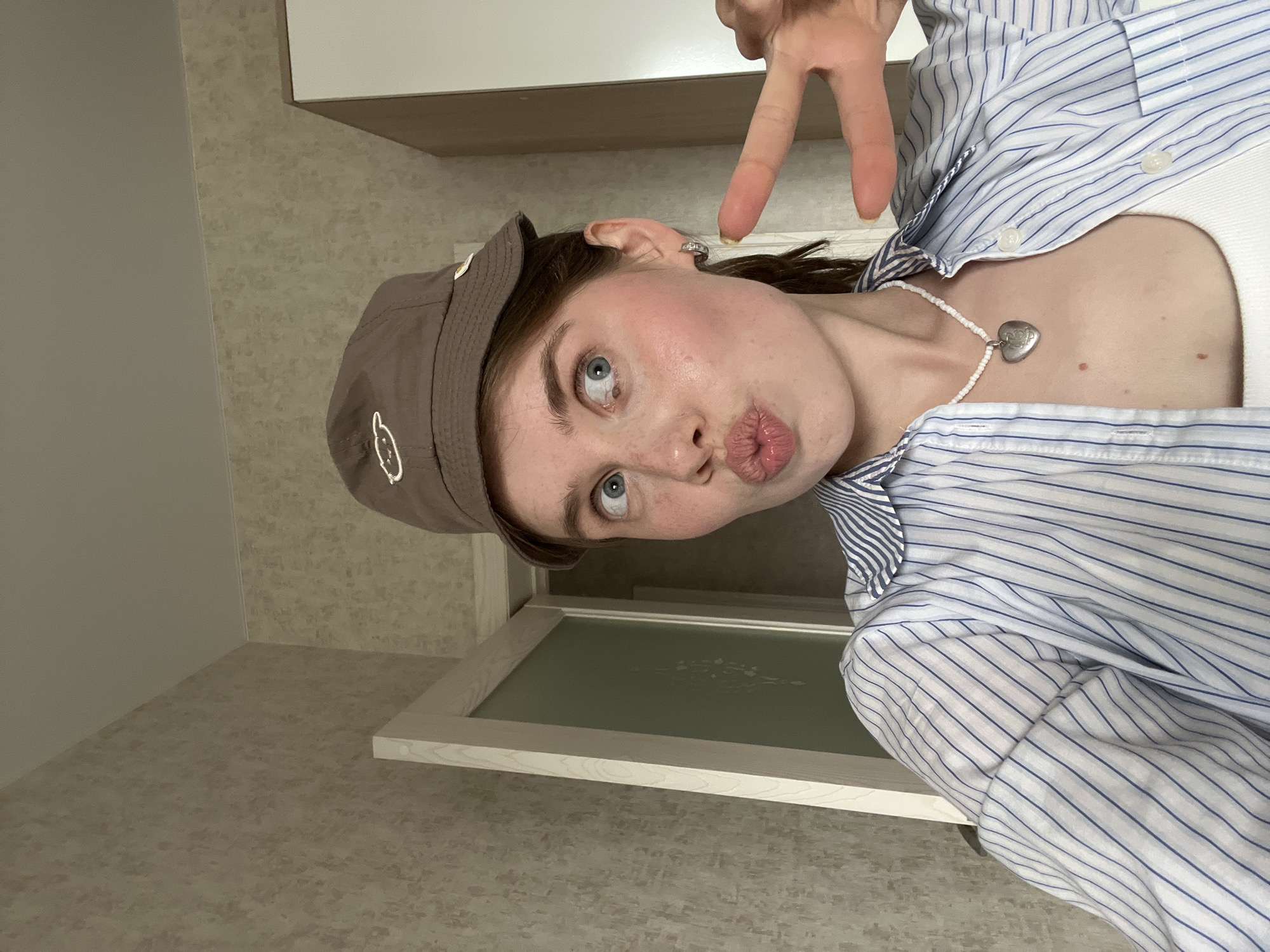 This image, situated in the city of Chaikovsky, Russia, features a young adult female captured in a close-up indoor selfie. She has fair skin with visible freckles on her nose and cheeks, and her blue eyes are wide and directed towards the upper left from the viewer's perspective, appearing slightly crossed. Her lips are pursed in an exaggerated "kissing" expression.
Her brown hair is pulled back, with some strands visible around her face. She wears a brown bucket hat tilted backward, adorned with a simple white outline of a cloud or sheep-like shape embroidered on the front. A small, silver-toned hoop earring is visible in her right ear. Her attire consists of a white tank top layered under a blue and white vertically striped button-down shirt, worn unbuttoned. Around her neck is a white beaded necklace with a prominent silver-toned heart-shaped locket pendant.
Her left hand is raised, presenting a "V" or "peace" sign with the index and middle fingers extended. A yellowish substance is discernible on the fingernail of the extended index finger. The background reveals an interior room with light beige or brown textured wallpaper on the walls and a plain light-colored ceiling. A partially open door with a light-colored frame and a frosted or light green glass insert is visible behind her left shoulder. To the far right, a corner of a white cabinet or wardrobe is present. The lighting appears to be ambient.