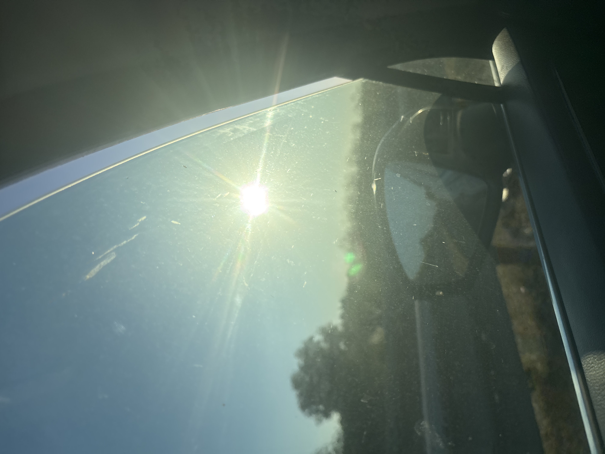 The image is a view from inside a vehicle, looking out the passenger side window. The sun is shining brightly, creating lens flare and streaks across the glass, which is also marked with smudges and dust. The sky is a clear blue. In the distance, silhouetted trees and vegetation are visible, suggesting a rural or suburban setting. A car's side-view mirror is partially visible in the foreground, reflecting the sky and some of the surrounding environment. The interior of the car, including the window frame and part of the dashboard or door panel, is also visible, rendered in dark tones. No specific landmarks indicating Burgas, Bulgaria, are present.