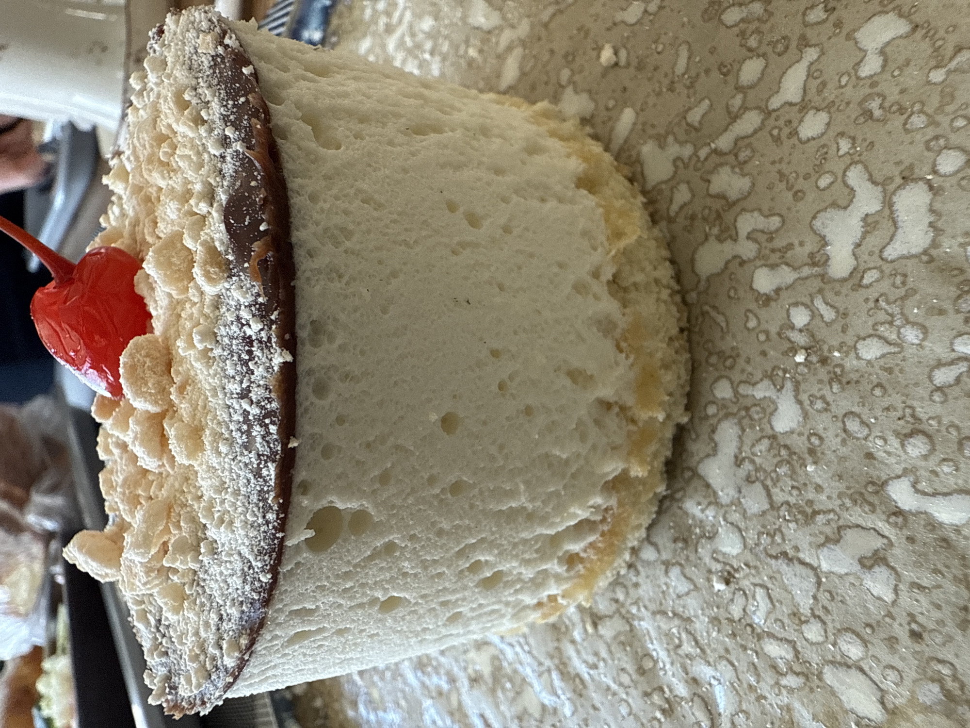 This close-up image, likely taken in Votkinsk, Russia, displays a section of a dessert or pastry item oriented vertically. The primary subject is a slice of a white, airy, porous substance resembling bread or sponge cake, which forms the bulk of the item. Along one edge, a thick, dark brown layer, appearing to be chocolate frosting or ganache, is visible. This chocolate layer, along with an adjacent section of the white base, is generously covered with light-colored, granular crumbs or powdered sugar, giving it a textured, frosted appearance.
A bright red maraschino cherry with its stem intact is prominently positioned on the crumb-covered side of the dessert, adding a pop of color. The background consists of a light brown or beige surface, possibly a tray or countertop, characterized by numerous irregular, lighter-colored splotches and rings, resembling dried liquid marks or a distinct pattern. The overall lighting is bright and even, highlighting the textures of the food item. No specific activity is taking place, but the item appears to be prepared and presented, possibly for consumption or display. There is no visible text in the image.