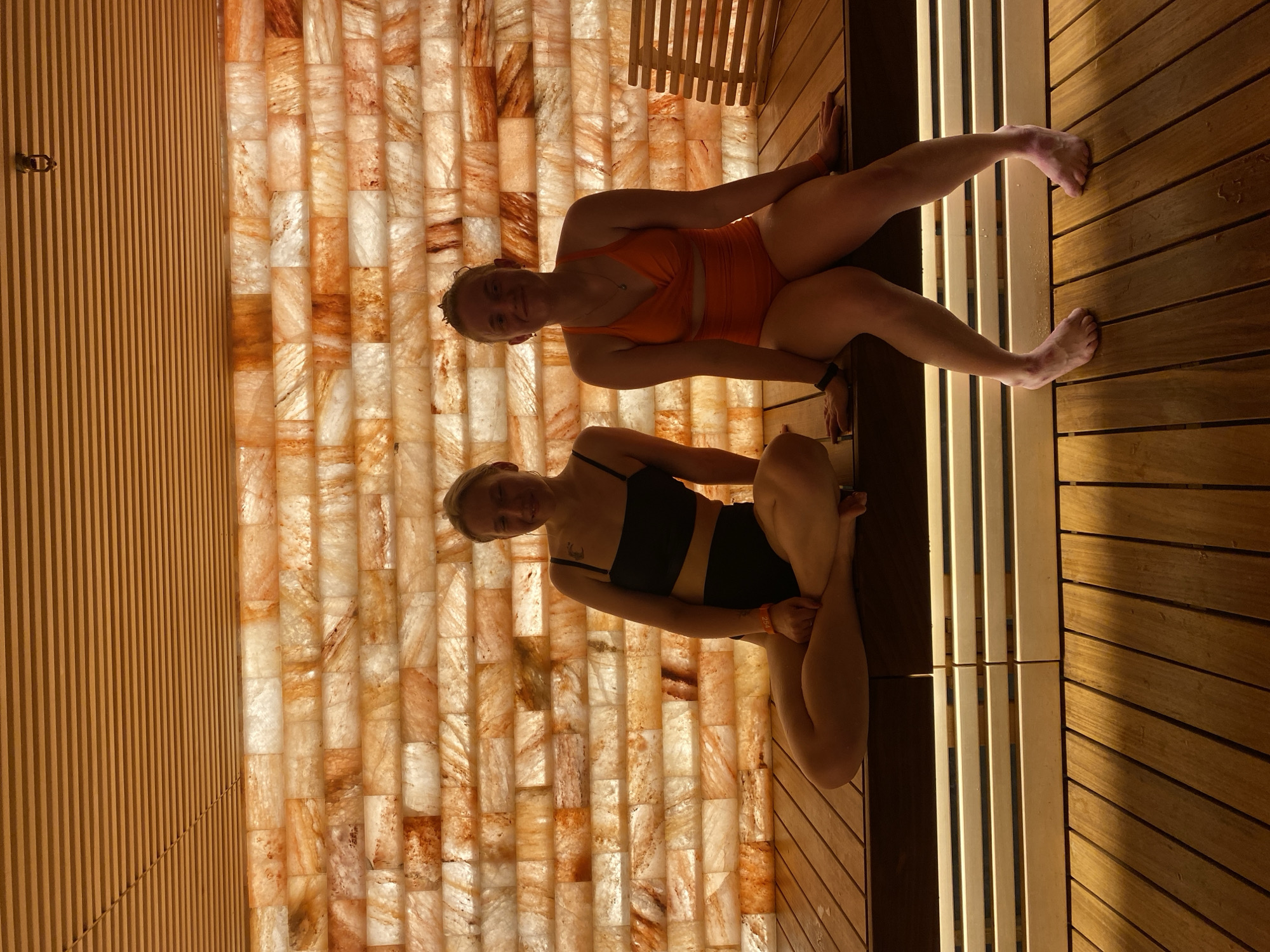 Two Caucasian women are sitting in a room constructed of Himalayan salt bricks and wood. One woman wears an orange swimsuit, and the other a black one. The location of the scene in Votkinsk, Russia is not apparent in this image.