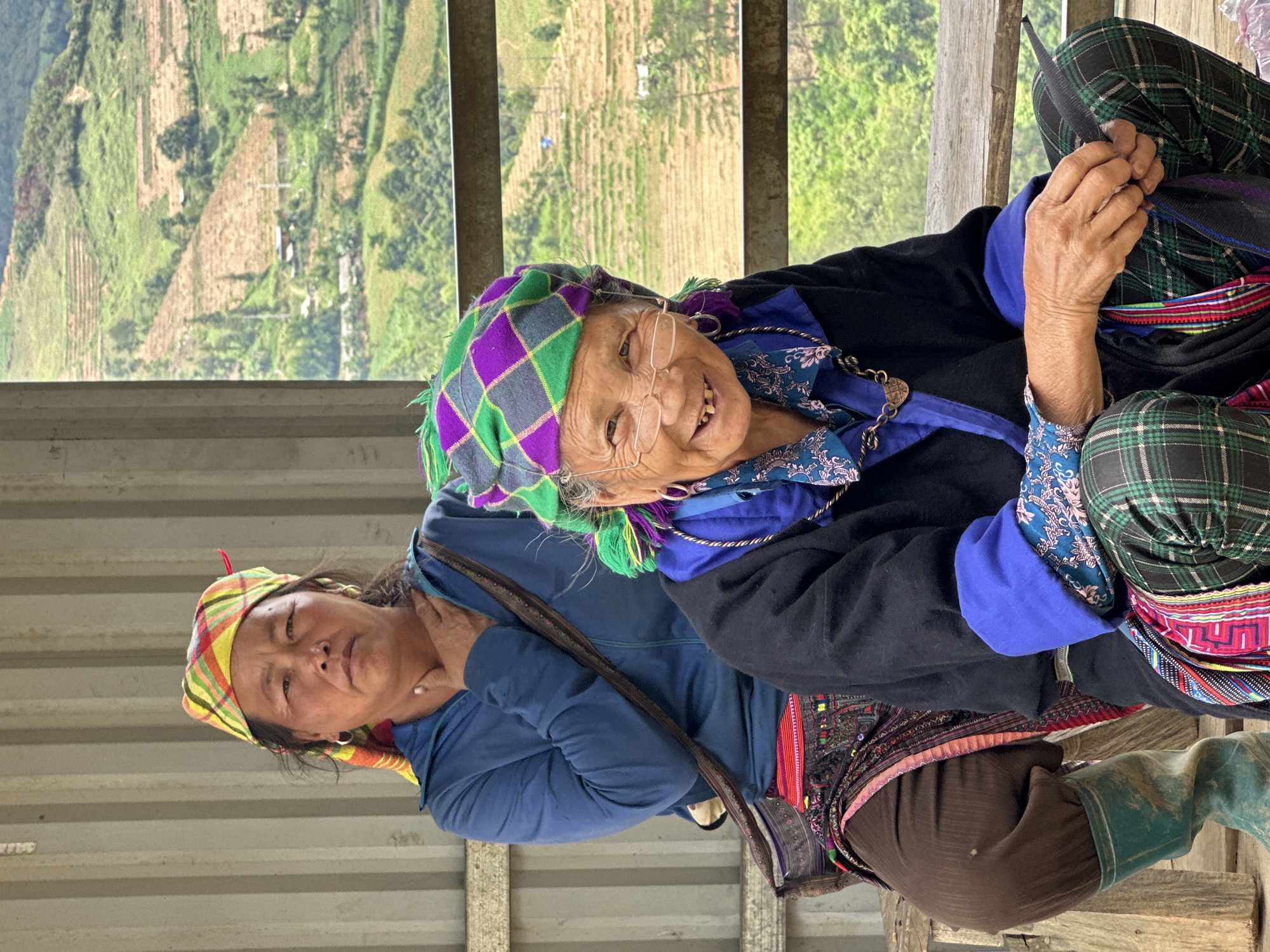 Two women are seated in a sheltered outdoor area in Dế Xu Phình, Vietnam, with a mountainous, terraced agricultural landscape visible in the background.
The elderly woman in the foreground is seated, smiling and looking directly at the viewer. She wears spectacles, a green and purple plaid headwrap with tassels, a black long-sleeved jacket with blue cuffs, and a blue patterned shirt underneath. Her lower attire consists of green and black plaid pants, adorned with colorful patterned sashes. She wears silver hoop earrings and a copper-toned necklace with a circular pendant. Her hands are clasped in her lap, holding a dark strap.
Behind her, the second woman, appearing middle-aged, sits with a neutral expression, also looking forward. She wears a yellow, red, and green plaid headwrap and a blue long-sleeved zippered jacket. Over her brown pants, she wears a dark patterned skirt or apron with colorful embroidery. Her right hand rests on her collarbone, and a dark shoulder bag with a patterned strap is visible across her body. She wears silver hoop earrings and mud-stained teal or green rubber boots.
The immediate background behind the women includes a corrugated metal wall to the left and a horizontal metal railing to the right. Beyond the railing, the distant landscape features verdant hills, extensive terraced fields, scattered trees, and small, distant structures under bright daylight. The women are seated on a wooden bench or platform.