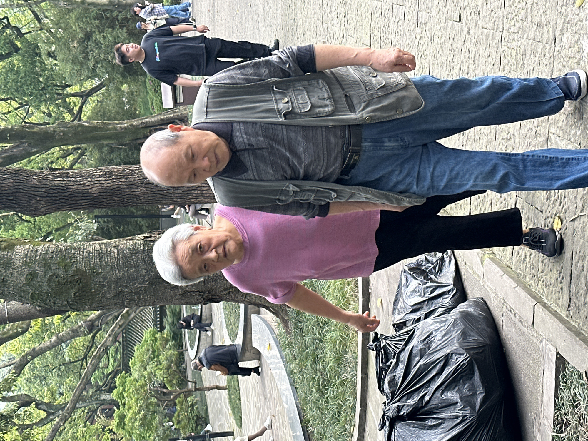 The image captures a scene in Yuzhong District, China, featuring an elderly couple standing on a paved walkway. The woman, wearing a pink sweater and black pants, stands closer to the camera, while the man, dressed in jeans, a grey vest, and a striped shirt, stands slightly behind her. Two large black trash bags sit beside them. A tree trunk partially obscures the view between the couple. In the background, a younger man in a black shirt and pants is visible, along with other individuals walking along the walkway. The environment includes trees, greenery, and a sculpted concrete element. The overall atmosphere suggests a public park or garden setting.