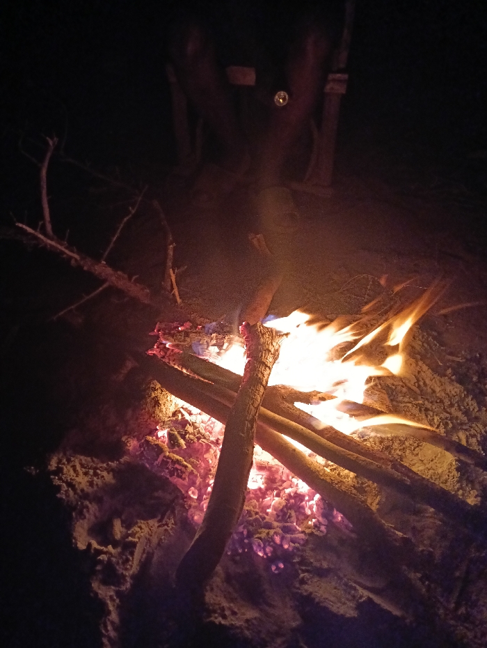 The image captures a bonfire burning at night in an outdoor setting in Kyamatu, Kenya. The central focus is a pile of wooden logs and branches actively consumed by fire, displaying bright yellow, orange, and red flames. Beneath the flames, a substantial bed of glowing red and purple embers is visible. The ground immediately surrounding the fire is light-colored and granular, consistent with sand or loose dirt.
The background is largely obscured by deep shadows, indicating nighttime conditions. In the upper central portion of the image, a dark, indistinct vertical mass is present, appearing to be a large, upright object or structure, possibly a tree trunk or a post. Above this mass, a small, distinct circular reflection or point of light is discernible. No identifiable individuals or specific human actions are evident within the frame, apart from the implied presence required to start and maintain the fire. The primary interaction depicted is the combustion of wood.