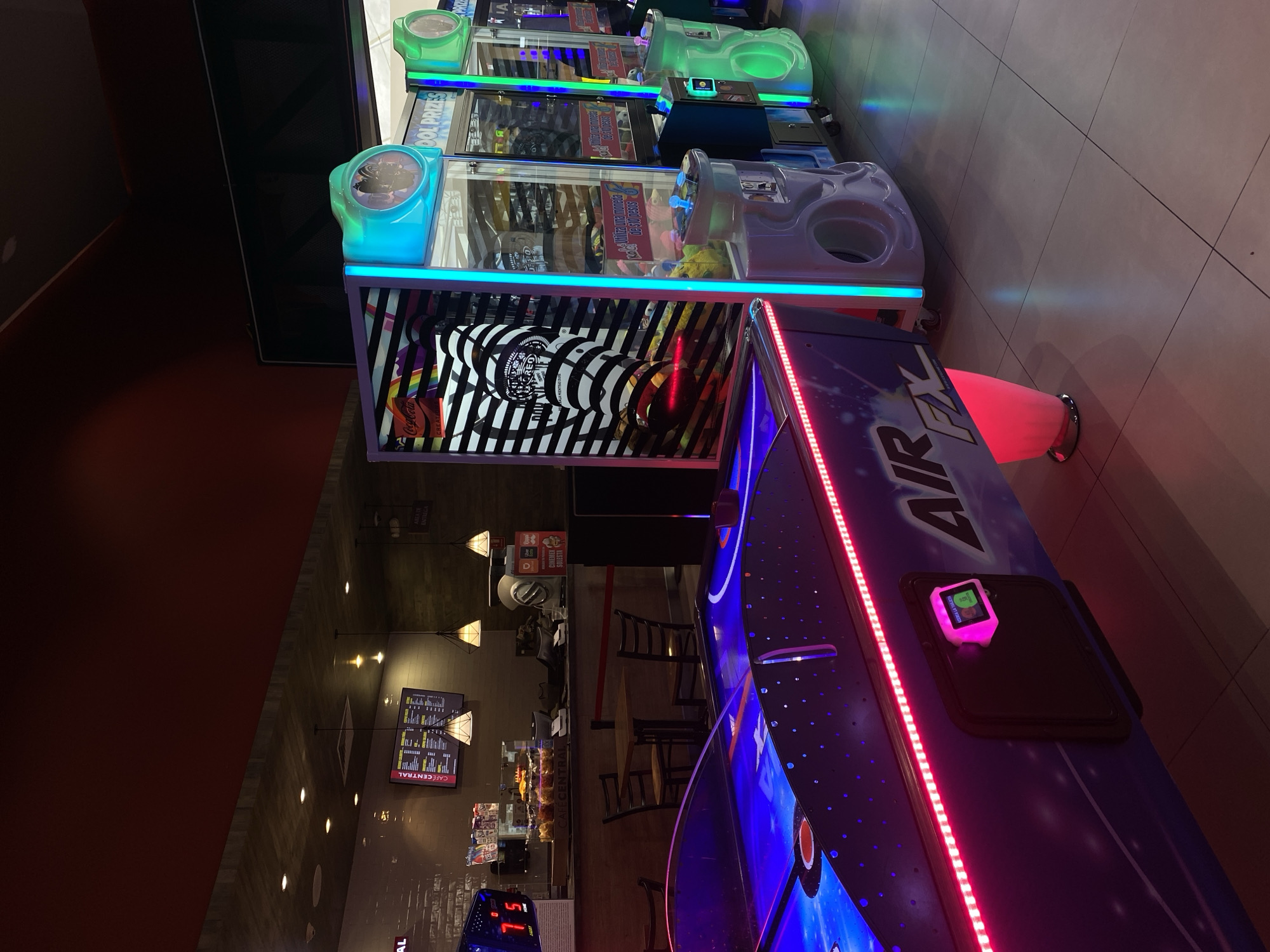 The image captures an indoor entertainment and dining space, likely an arcade connected to a cafe, illuminated by artificial light, suggesting an evening or night setting.
In the foreground, a prominent blue and purple air hockey table, labeled "AIR PIX," glows with vibrant pink/red LED strips along its edge. A pink-illuminated card reader on the table displays "ACERCA LA TARJETA" (bring the card closer) and a score of "15." Behind it, a series of arcade machines, primarily claw games, are lit with bright green and blue neon lights. These machines showcase various prizes, including "Coca Cola" and "OREO" products, with signs like "COOL PRIZES" and "PRIZE" visible. Text on one machine indicates "Utiliza una moneda de (X) pesos" (use a coin of X pesos).
To the left and in the background, a cafe area features multiple dark tables and chairs. Overhead, decorative pendant lights cast a warm glow. Menu boards on the wall display "CAFÉ CENTRAL" and list items. A sign in this area includes "CINEMEX SOLESTA" and "ENTREGA" (delivery/pick-up). The floor throughout the venue is composed of light-colored tiles, and a solid, dark reddish-brown wall runs along the left side of the frame. No people are present, creating a quiet atmosphere despite the colorful and inviting environment of the games and cafe.